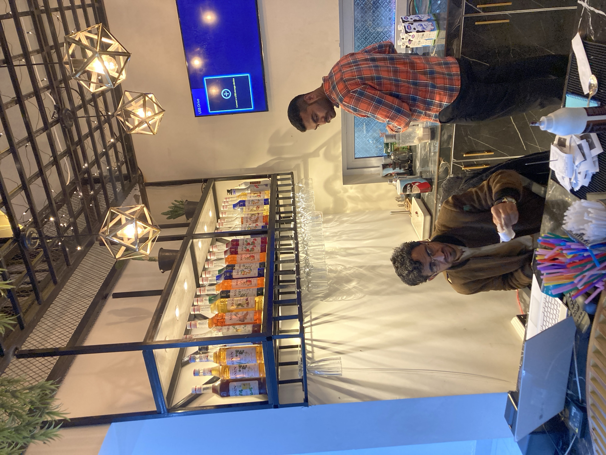 The image shows two men behind a bar with a variety of flavored syrups on shelves. The first man is wearing a red plaid shirt and standing behind the bar, while the second man is wearing a brown jacket and is looking at something in his hand, with a laptop open in front of him. A pile of colorful straws is on the counter to the right of the laptop. The location is likely a bar or restaurant. There's a flat-screen TV on the wall and a modern lighting fixture hanging from the ceiling. Bottles are displayed on shelves behind the men. The text in the image indicates a USB drive is connected. It's difficult to determine the city of Quetta City Tehsil, Pakistan with any certainty.