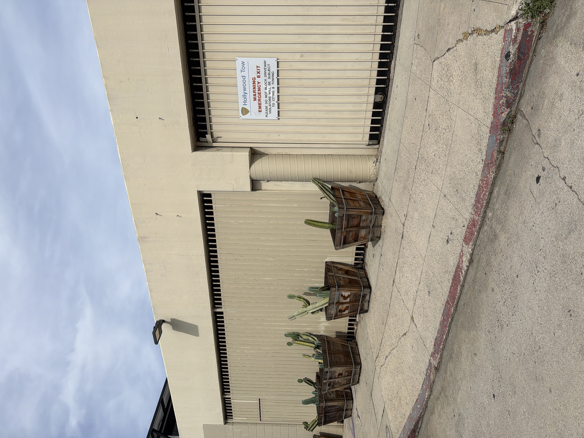 This outdoor daytime image displays the side of a light-colored building with a large metal gate, a concrete sidewalk, and several potted cacti. The perspective is tilted, showing a bright sky with some light cloud cover, indicating a sunny day.
The building's facade is a pale yellow or beige. A large, cream-colored metal roll-up gate or security barrier is prominently featured in the middle of the frame. Attached to this gate is a white rectangular sign that reads: "Hollywood Tow WARNING EMERGENCY EXIT PLEASE DO NOT BLOCK DRIVEWAY VIOLATORS WILL BE SUBJECT TO CITING & TOWING." Above the main gate, there's another barred opening, and a light fixture on the upper wall casts a clear shadow.
Along the base of the building, on the concrete sidewalk, are four large, square wooden planters. Each planter contains multiple green, columnar cacti of varying heights and angles. The concrete sidewalk itself is cracked in places, with some weeds growing through the fissures. A section of the curb visible in the foreground is painted red, typically signifying a no-parking zone.
No people are visible, and no specific activity is taking place. The "Hollywood Tow" sign and the presence of cacti consistent with arid climates strongly suggest a location in Los Angeles, United States.
