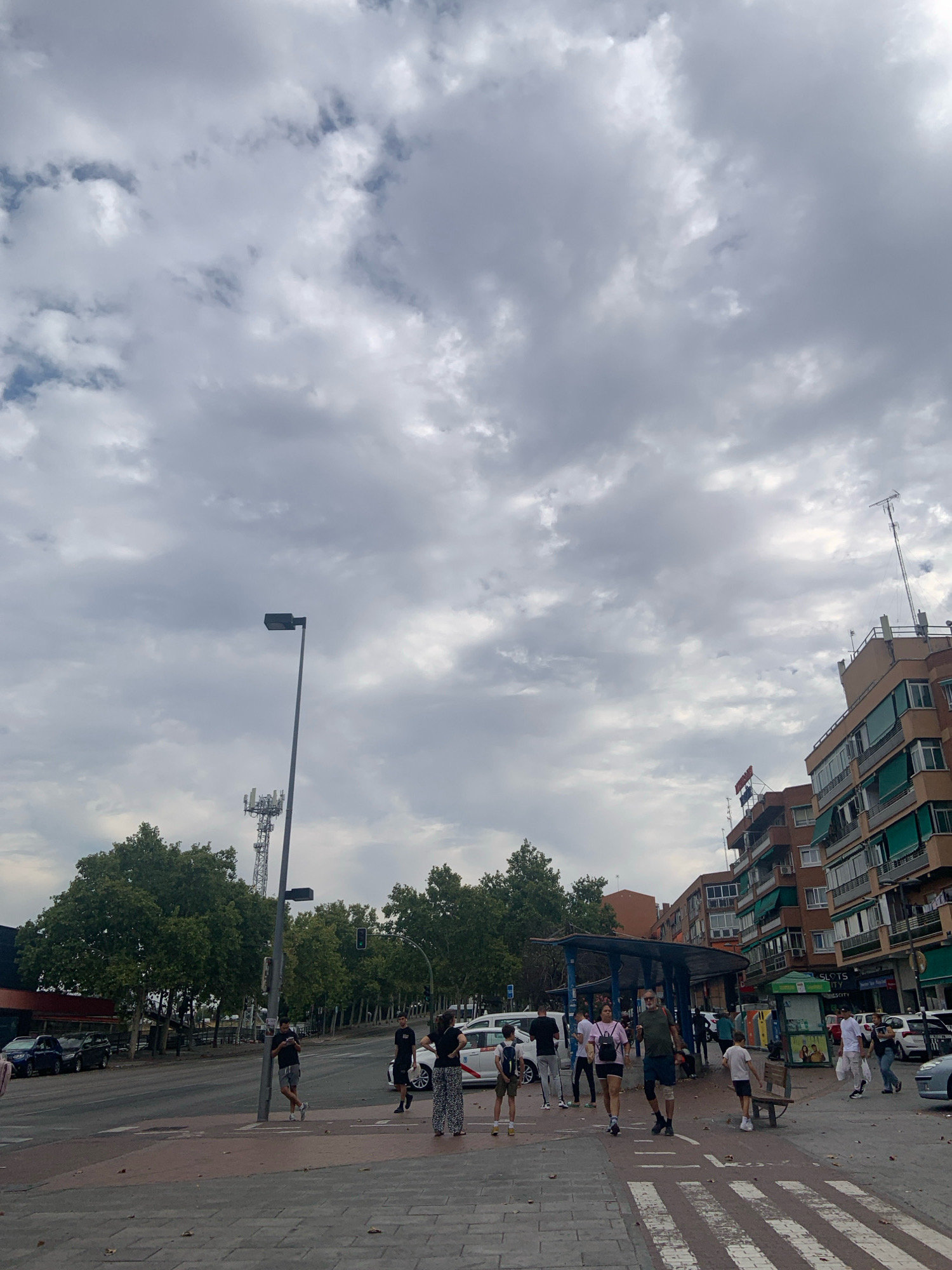 The image shows an urban scene in Alcorcón, Spain, under a cloudy sky. In the foreground, a pedestrian crossing is visible, leading to a sidewalk where several people are walking. Some individuals are casually dressed, while others appear to be engaged in conversations or observing their surroundings. A bus stop shelter is situated on the right side of the street, with a few people waiting or sheltering beneath it.
The background features a street with cars, including a white taxi. Tall, leafy trees line the street, providing some shade. Buildings with multiple stories, displaying various architectural styles and colors, dominate the backdrop. A tall communication tower and a street lamp are also prominent features in the distance. The overall atmosphere suggests a typical day in a Spanish city, with daily activities unfolding in a public space.