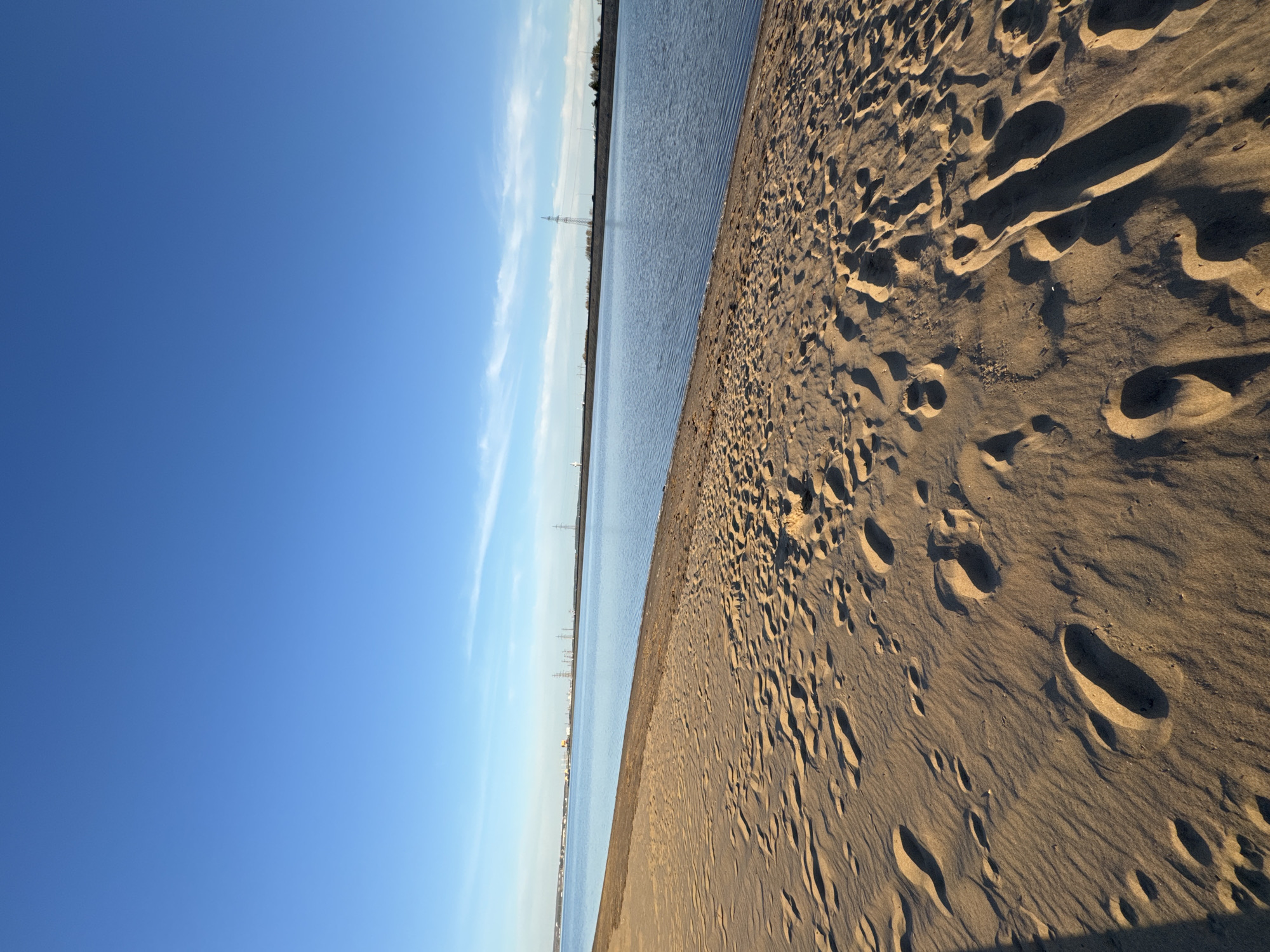 The image depicts a beach in Noviy, Russia. The right side of the frame is dominated by sand with numerous footprints. The left side features a body of water and a clear blue sky with wispy clouds. Utility poles are visible along the horizon.