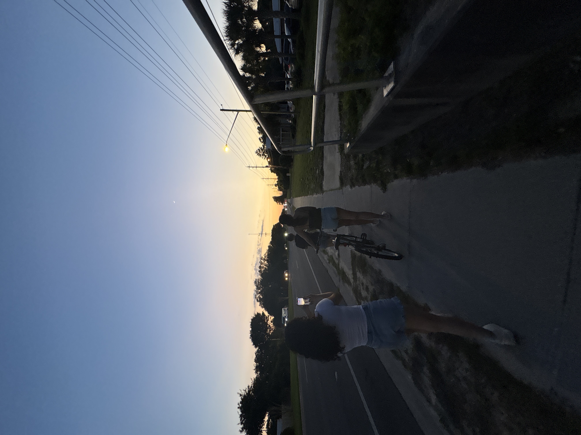 Here is a detailed description of the image:
**Scene:** The photograph appears to be taken on a paved path alongside a road. A metal railing runs parallel to the path. Trees and buildings are visible in the background. The sky is a gradient of blue to orange, suggesting either sunrise or sunset. A faint crescent moon is visible in the upper left.
**People:** Two individuals are on the path. One person is holding a phone and taking a photograph. The other person is standing with a bicycle.
**Objects:** A bicycle, a phone, a metal railing, trees, and buildings.
**Actions:** One person is taking a photo with a phone. The other person is standing with a bicycle.
**Interactions:** The individuals appear to be together, possibly enjoying a walk or bike ride.
**Environment:** The scene is set in a semi-urban environment with a combination of natural elements (trees) and constructed elements (road, buildings). The lighting suggests it is either early morning or late evening.
**Location:** Panama City Beach, United States of America. This is based on the general feel of the image and general information, as well as reference to other images in the same general location.
