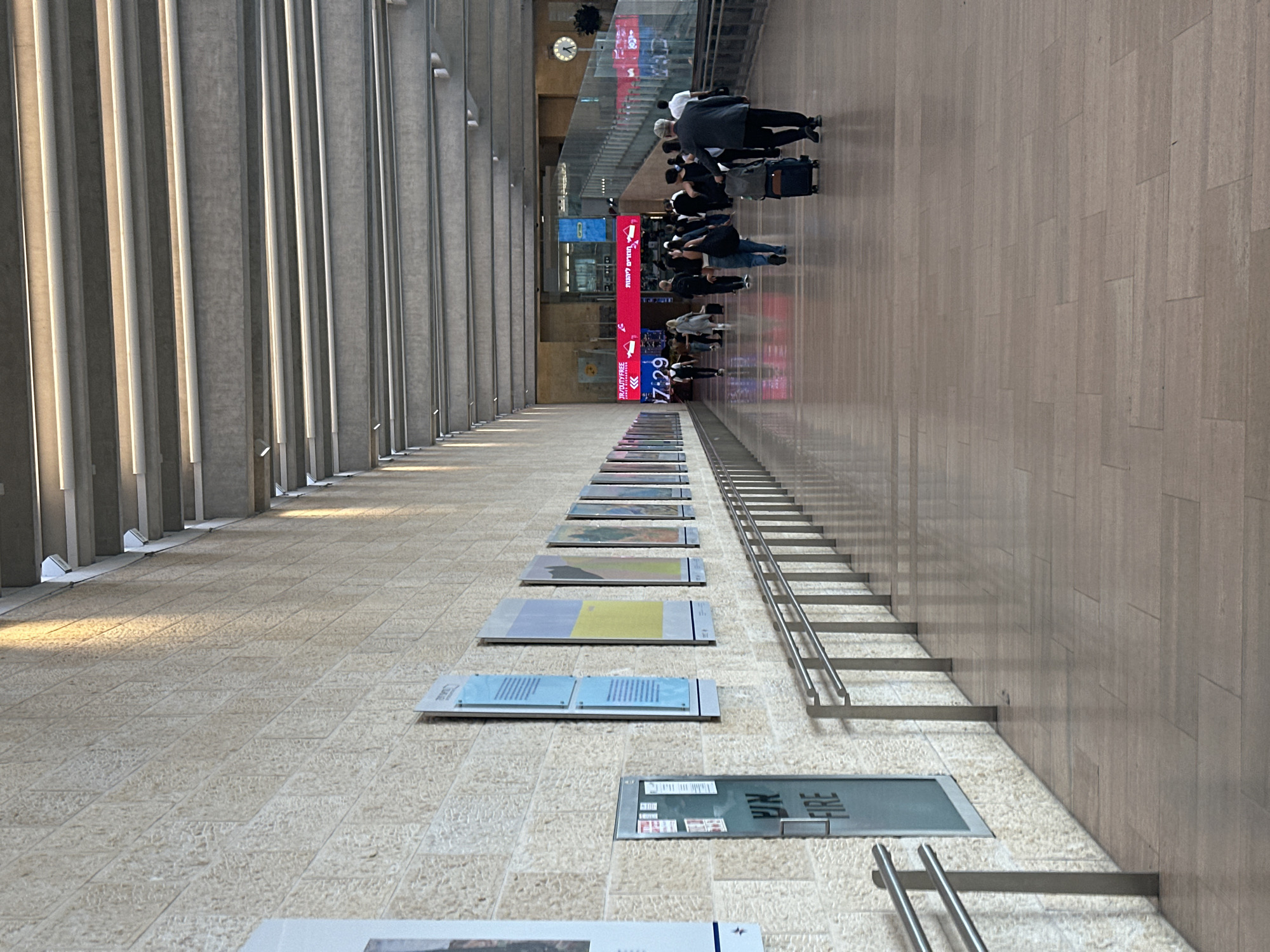 The image depicts a long, covered walkway within what appears to be an airport or a large public transportation hub in Tzafria, Israel. The walkway is lined with a textured, light-colored stone wall on the left, featuring several framed exhibits or posters. A prominent "FIRE" sign is visible on a metallic door on the left wall. On the right side of the walkway, a polished wooden floor reflects the overhead lighting and the activity within the space. A metal handrail runs along the length of the walkway.
In the distance, a crowd of people is seen walking, many with luggage, indicating travel. Digital signs displaying advertisements and information are visible in the background, including a "JR/DUTY FREE" sign and a clock. The ceiling is characterized by a modern design with exposed structural beams and integrated lighting. The overall atmosphere is one of transit and a busy public space.