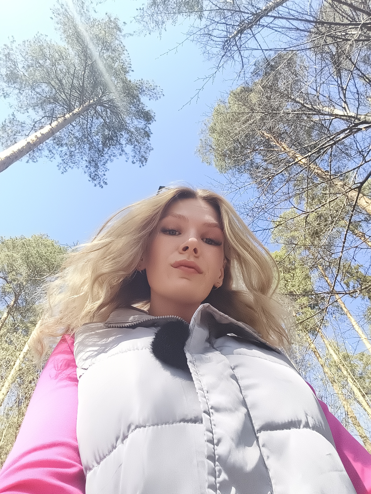 A low-angle, wide shot features a blonde woman outdoors, likely in a forest, with a clear blue sky visible through the trees. The woman is wearing a pink top and a puffy, light grey vest. The background consists of tall trees, suggesting a natural environment in Izhevsk, Russia. Bare branches are interspersed with evergreen foliage. The sun seems to be shining from above.