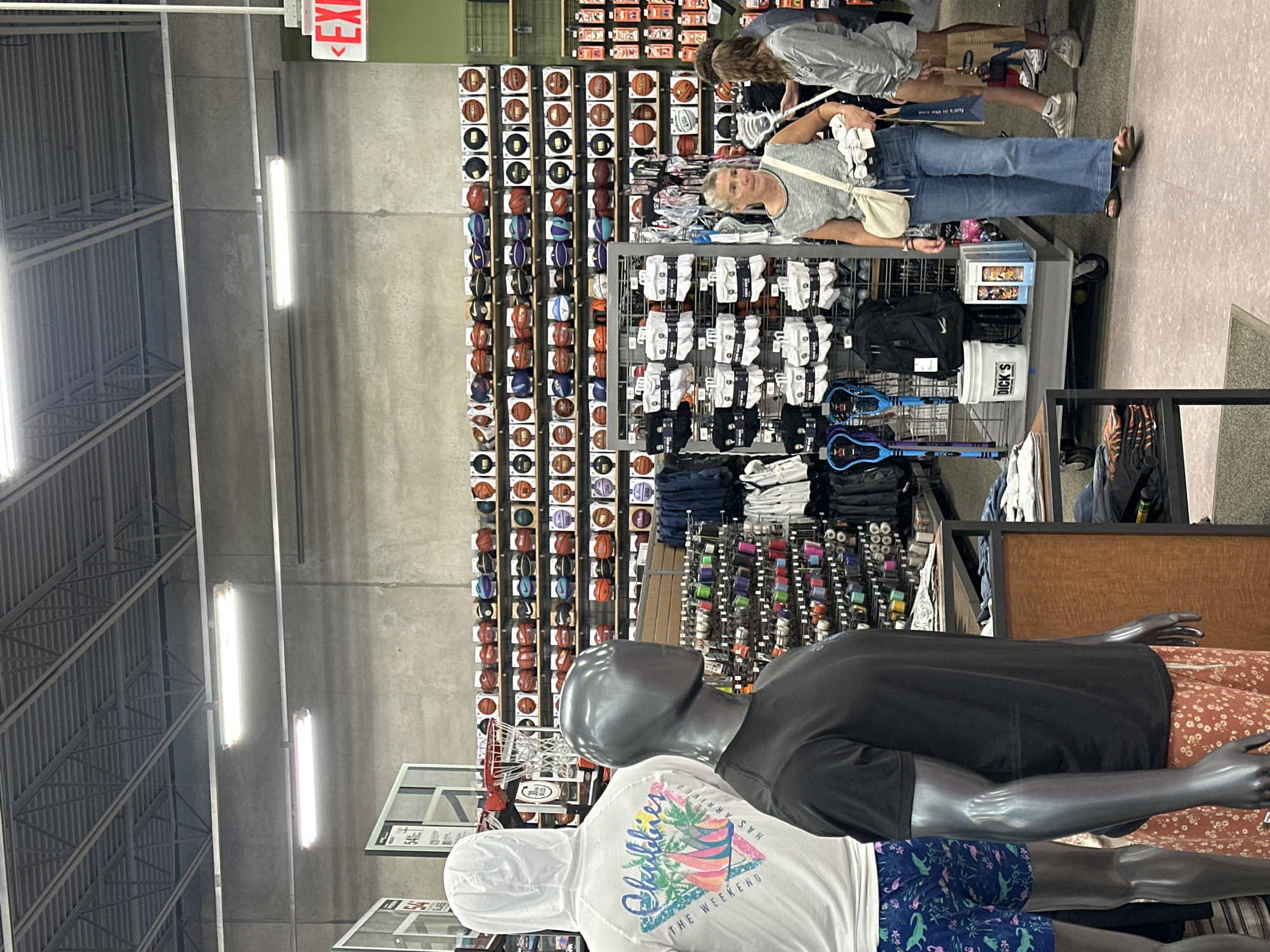 In a retail sporting goods store, likely Dick's Sporting Goods in Algonquin, Illinois, a variety of merchandise is displayed. The background features shelves stacked with basketballs, and various sports accessories such as socks and bags are visible on display racks. In the foreground, two mannequins are dressed in athletic wear. One mannequin wears a white hooded shirt with a graphic print and patterned shorts, while the other is dressed in a black t-shirt and patterned shorts. Three women are present in the store. One woman in the mid-ground, wearing jeans and a striped top, is looking towards the left side of the image. Another woman, wearing a gray sweatshirt and shorts, is standing to the right of the first woman, holding a shopping bag. A third woman is partially visible to the right of the second woman, also holding a shopping bag. The overall environment is that of a brightly lit retail space with an industrial ceiling.