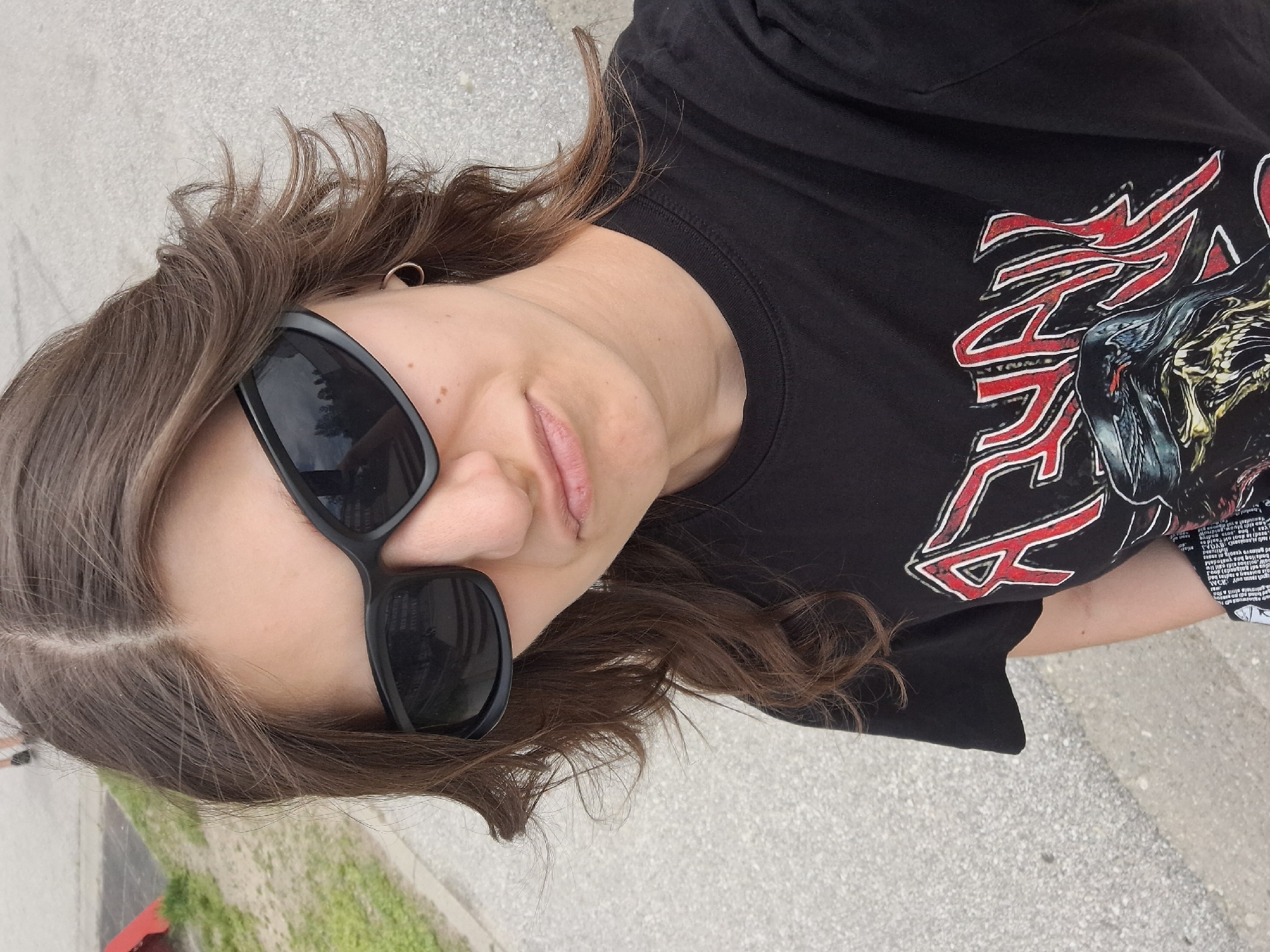 A person with medium-length brown hair is wearing black sunglasses and a black T-shirt with a red and silver design. The location appears to be outdoors on a paved surface with greenery visible in the background. The specific location in Panagyurishte, Bulgaria, cannot be determined from the image.