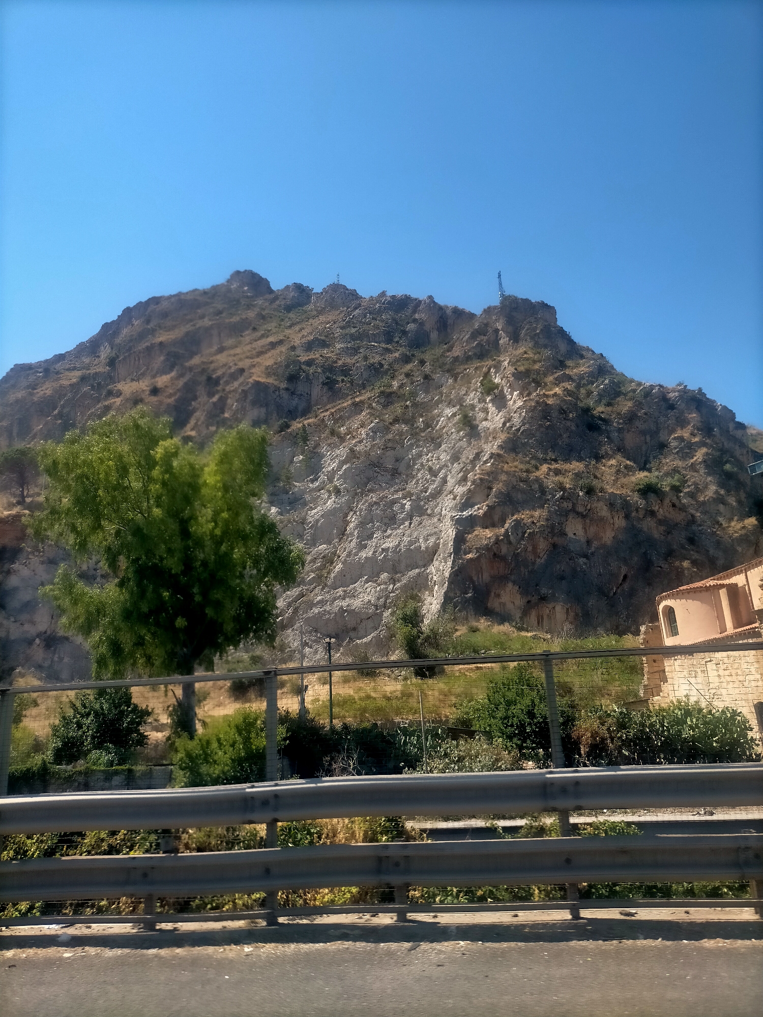 A bright, clear blue sky dominates the upper portion of the image. Below the sky, a large, rocky mountain occupies the majority of the frame, its slopes covered with sparse, dry vegetation and a prominent, lighter-colored rock face, suggesting a quarry or erosion. A tall, green tree stands on the left side of the mountain, its leaves appearing lush. A metal guardrail stretches across the foreground, obscuring the lower part of the scene. Behind the guardrail, a fence encloses an area with more vegetation, including bushes and some greenery. On the right side of the image, a portion of a building with a reddish-orange roof and beige stone walls is visible, suggesting it is an older structure. The overall impression is a landscape with a rugged natural element juxtaposed with human infrastructure and architecture. The image appears to be taken from a moving vehicle on a road, given the slight blur and the perspective of the guardrail. The location is specified as being in Palermo, Italy.