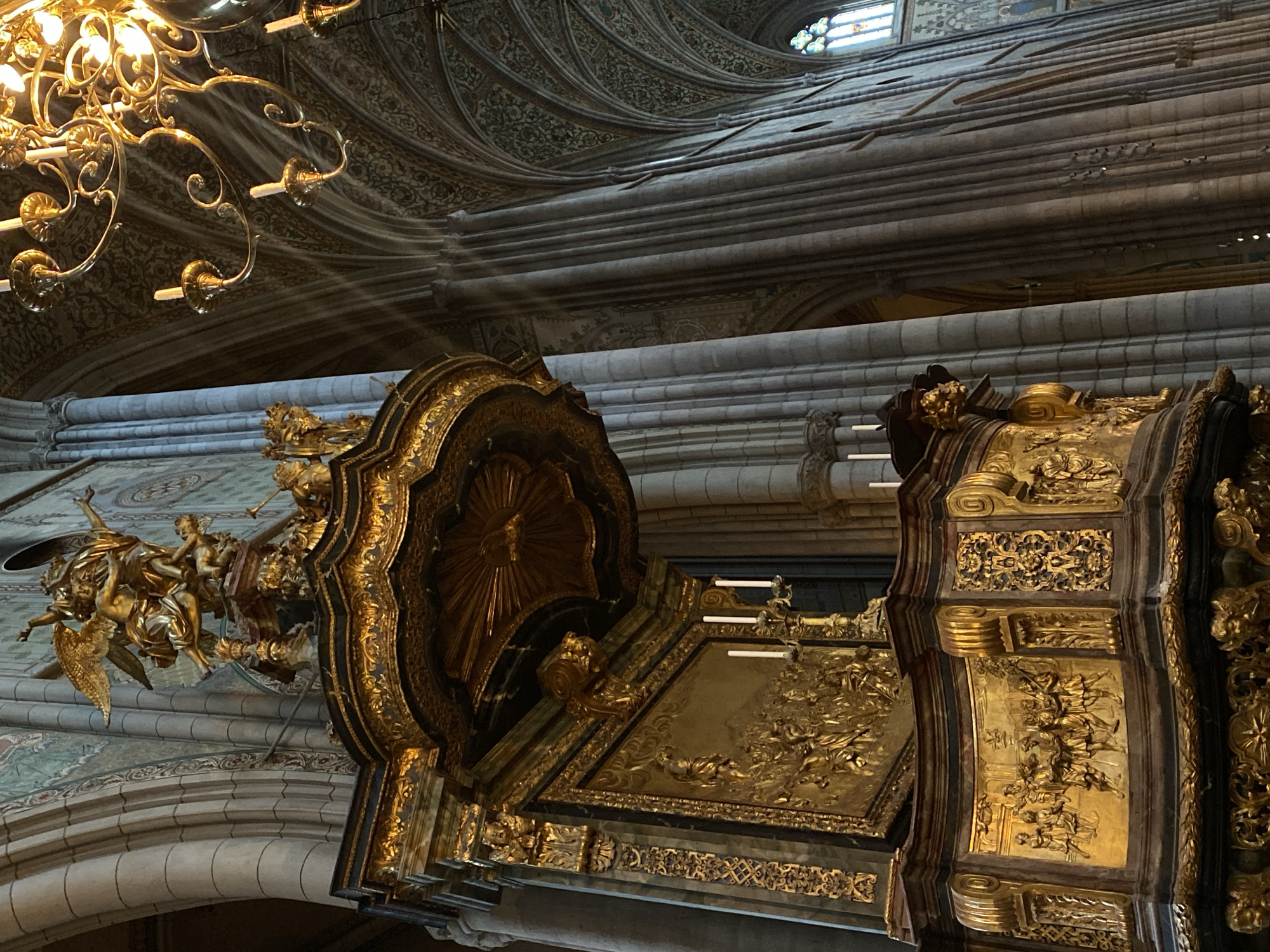 The image depicts the interior of Uppsala Cathedral in Uppsala, Sweden. A large, ornate gold reliquary or shrine is prominently featured. The reliquary has panels decorated with relief carvings. To the left is a sculpture of angels. A chandelier hangs from the ceiling in the upper left corner. The ceiling has an intricate design, and light streams into the building.