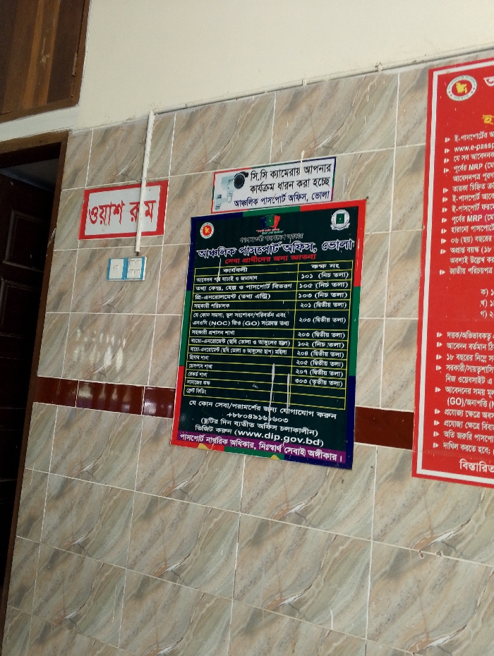 The scene is an interior wall of a building, likely a government office, in Bhola, Bangladesh. The wall is tiled with beige-brown tiles. On the wall are two posters with text in Bengali. The poster on the left is a white poster with red lettering at the top that says "জরুরী ফোন" and a yellow star. Below the star is a table in Bengali that lists various services and their corresponding fees. The poster on the right is red with white lettering. The lettering includes a website address for the "e-passport," contact information, and a list of different fees. The poster on the left is slightly torn. The scene is a government office. The posters appear to be instructions for applying for various government services. There is a doorway in the background that has a brown door. There is also a light switch to the left of the posters. There is no one in the image.