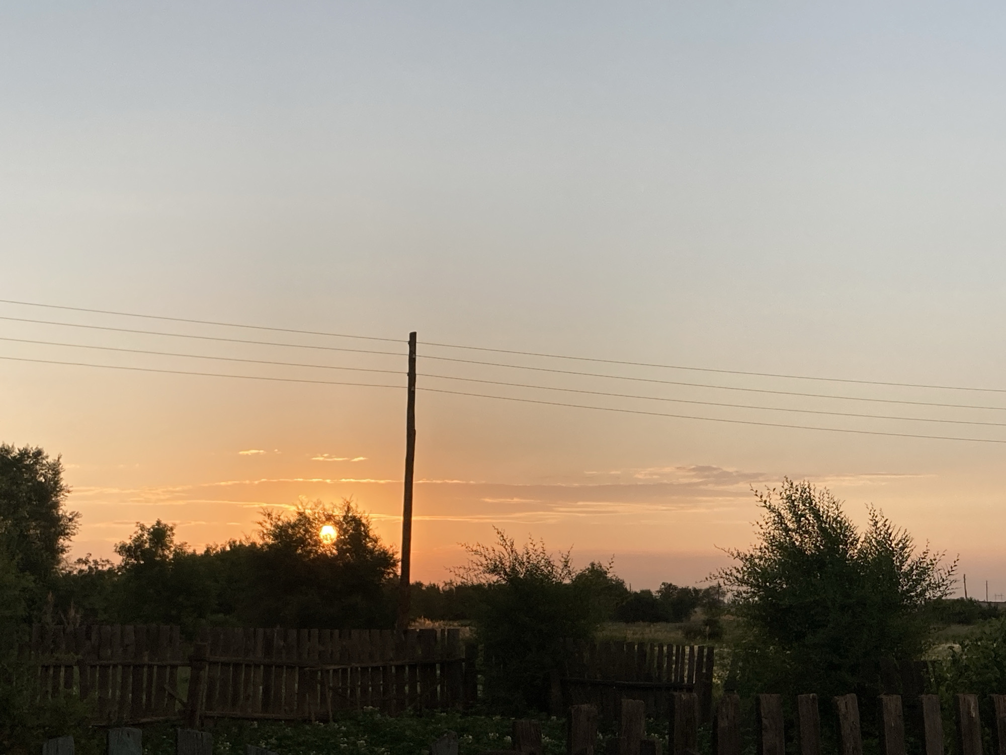 This media file depicts a serene rural landscape at sunset in Rakity, Russia. The scene is dominated by a pale orange and yellow sky, transitioning to a light blue at the top, indicating the end of the day. The sun, a bright orb, is partially obscured by silhouetted trees on the horizon, casting a warm glow and creating a peaceful, tranquil atmosphere.
In the foreground, a rustic wooden fence runs horizontally across the frame, suggesting a property boundary or the edge of a garden. Beyond the fence, lush green foliage and trees create a dense, natural backdrop. A solitary utility pole with several wires stretching across the sky stands to the left of the frame, a man-made element contrasting with the natural beauty of the setting. The overall impression is one of quiet and simplicity, characteristic of rural life. There is no visible text in the image.