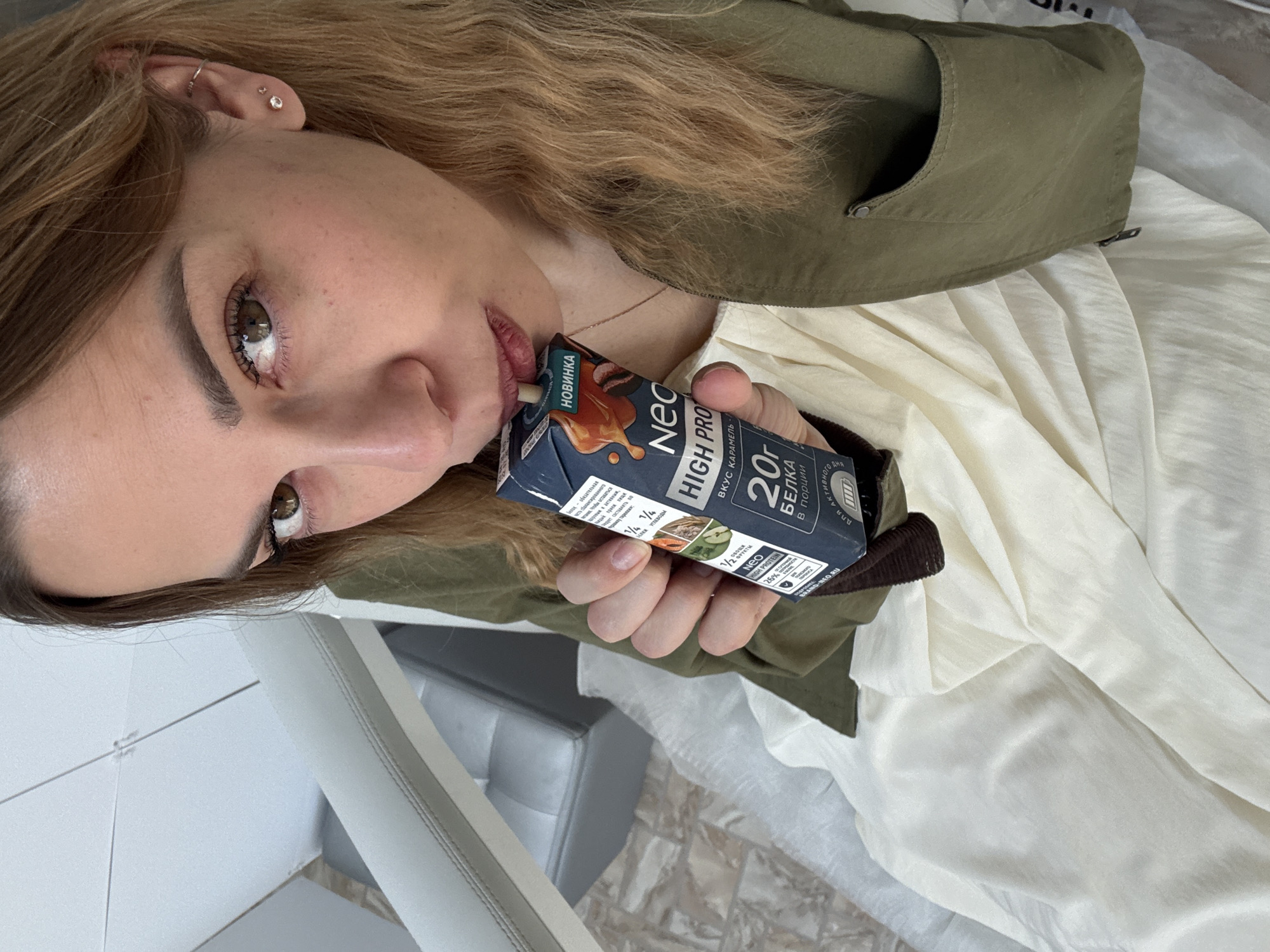 A young woman is drinking a "Neo High Pro" protein drink. The drink is caramel-flavored and contains 20g of protein per serving. The woman is wearing a green jacket and a light-colored shirt. Her hair is light brown. The location is likely Ust-Ilimsk, Russia, based on text on the package.