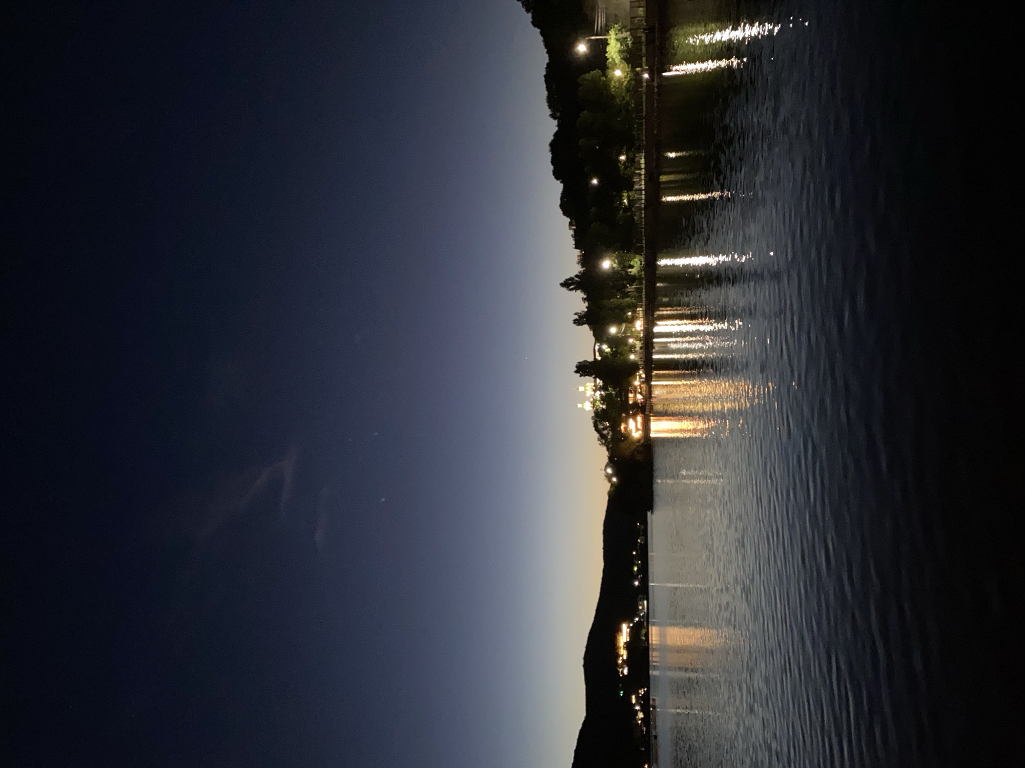 The image captures a nighttime view of a body of water in Abrau-Dyurso, Russia, likely during twilight or after sunset given the deep blue of the sky. The water is calm, reflecting the lights from the town visible on the shoreline. The town appears to be situated on rolling hills, with illuminated buildings and streetlights dotting the landscape. Dense trees line the shore in some areas, with their silhouettes dark against the illuminated background. The water itself is dark, with the reflections of the lights creating streaks of yellow and white on its surface. The scene conveys a peaceful, serene atmosphere characteristic of a waterfront town at night.