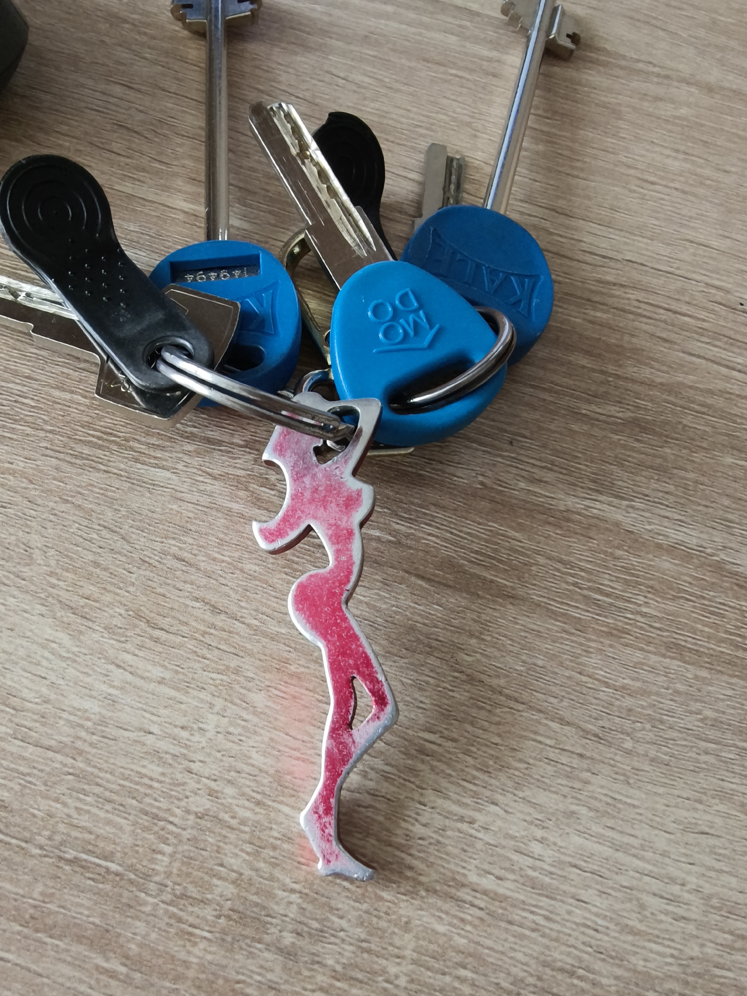 A cluster of keys and a distinctive keychain are resting on a light brown, wood-grained surface, likely a table or floor. The lighting suggests an indoor setting during daytime.
The keys include several long, silver-colored metal keys, some shorter, darker metal keys, and a key with a black plastic head. Attached to the key ring are three round or oval blue plastic key fobs.
The most prominent feature is a unique keychain shaped like a stylized female silhouette. It has a worn appearance, predominantly red with areas where the underlying silver material is visible.
Visible text can be observed on the blue key fobs, which are oriented upside down in the image. One fob clearly displays the text "MO DO". Another fob shows "KALE". A third fob has a sequence of numbers, "149494".
The scene is static, capturing the keys laid out, with no discernible activity or specific events taking place. The location context provided is Kryvyi Rih, Ukraine.