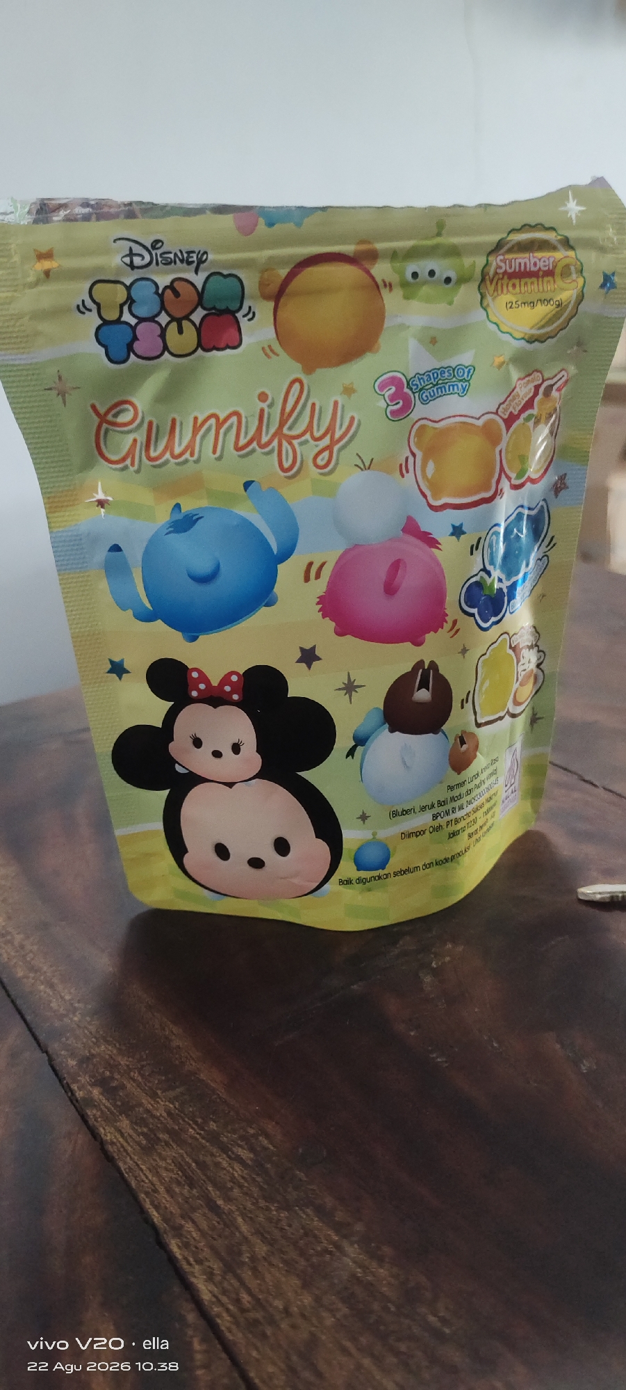 The image is a close-up, eye-level, slightly overhead shot of a package of Disney Tsum Tsum Gumify gummies. The packaging is primarily yellow and features various Tsum Tsum characters, including Winnie the Pooh, Mickey Mouse, Minnie Mouse, Stitch, and an alien from Toy Story. The text on the package is in English and Indonesian and indicates that the gummies are a source of Vitamin C. The package is placed on a dark wooden surface, likely a table. The lighting is bright and appears to be from overhead. The background is out of focus and appears to be a white wall. The image does not show the city of Simo, Indonesia, or any people.