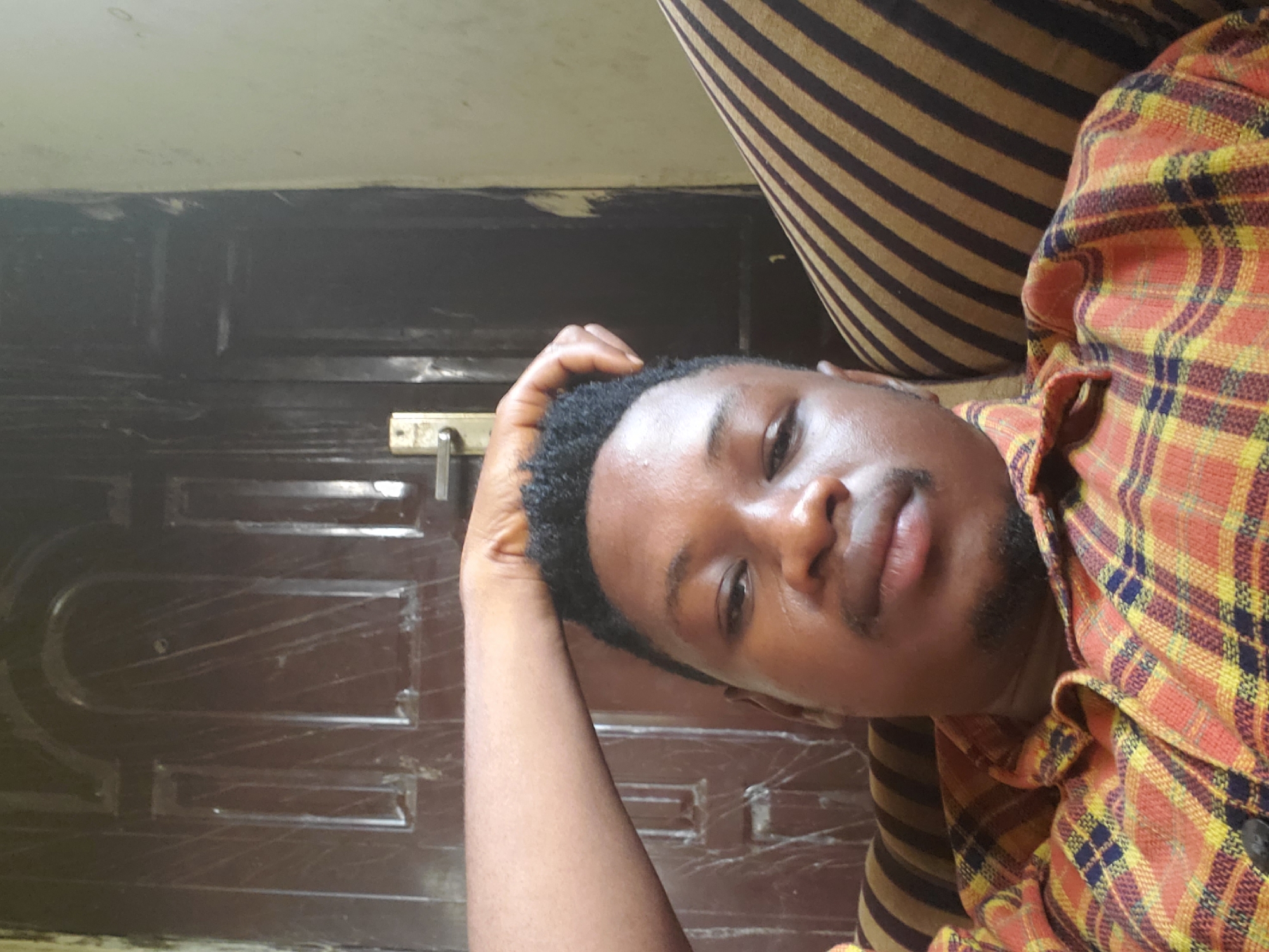 A man lies on a striped surface, wearing a plaid shirt. He is positioned in front of a brown door with silver hardware. The location is Mararaba, Nigeria.