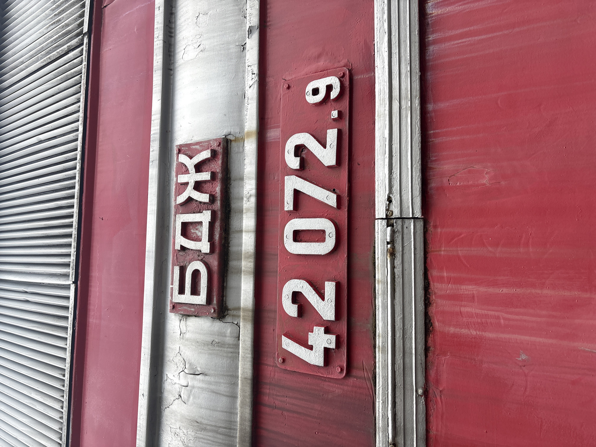 The image shows a close-up view of the exterior of a freight train car in Sofia, Bulgaria. The car is painted a faded red with silver accents. Two raised, painted plates are affixed to the side of the car. The plate on the left displays the Cyrillic letters "ВДЖ" vertically. The plate on the right displays the numbers "42072.9" vertically. The plates are likely associated with Bulgarian State Railways (BDZ), a state-owned railway company in Bulgaria. To the left of the train car is a metal grate.