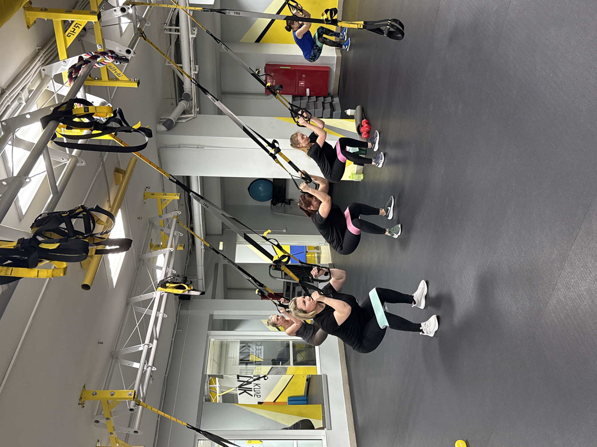 The image shows a group of women exercising in a fitness studio. The studio is located in Chaikovsky, Russia. The women are using TRX suspension training equipment. They are wearing athletic clothing and are focused on their workout. The studio has a black floor and a yellow and white color scheme. There are windows and mirrors along one wall. Other fitness equipment is visible in the background.