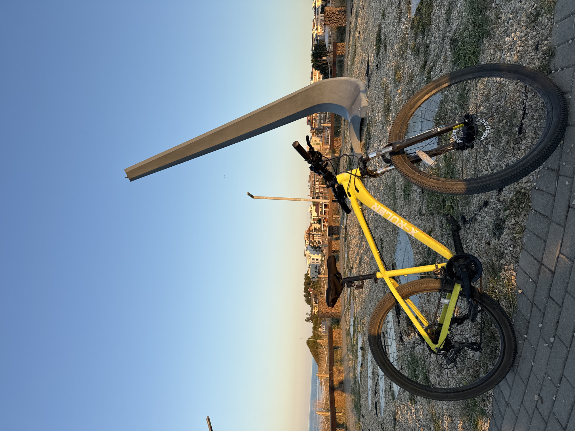 A bright yellow bicycle stands on a gravel and paved surface in Lozenets, Bulgaria, with a clear blue sky overhead. In the background, the sea is visible, and beyond that, a town with buildings of various colors is nestled against a hillside. A large, abstract concrete sculpture with a curved, angular form is positioned behind the bicycle, dominating the middle ground. The scene appears to be a public space, possibly a promenade or park, given the presence of benches and low wooden fencing lining the path leading towards the sea. The lighting suggests it is late afternoon or early evening, with warm sunlight casting long shadows. There are no people visible in the image.