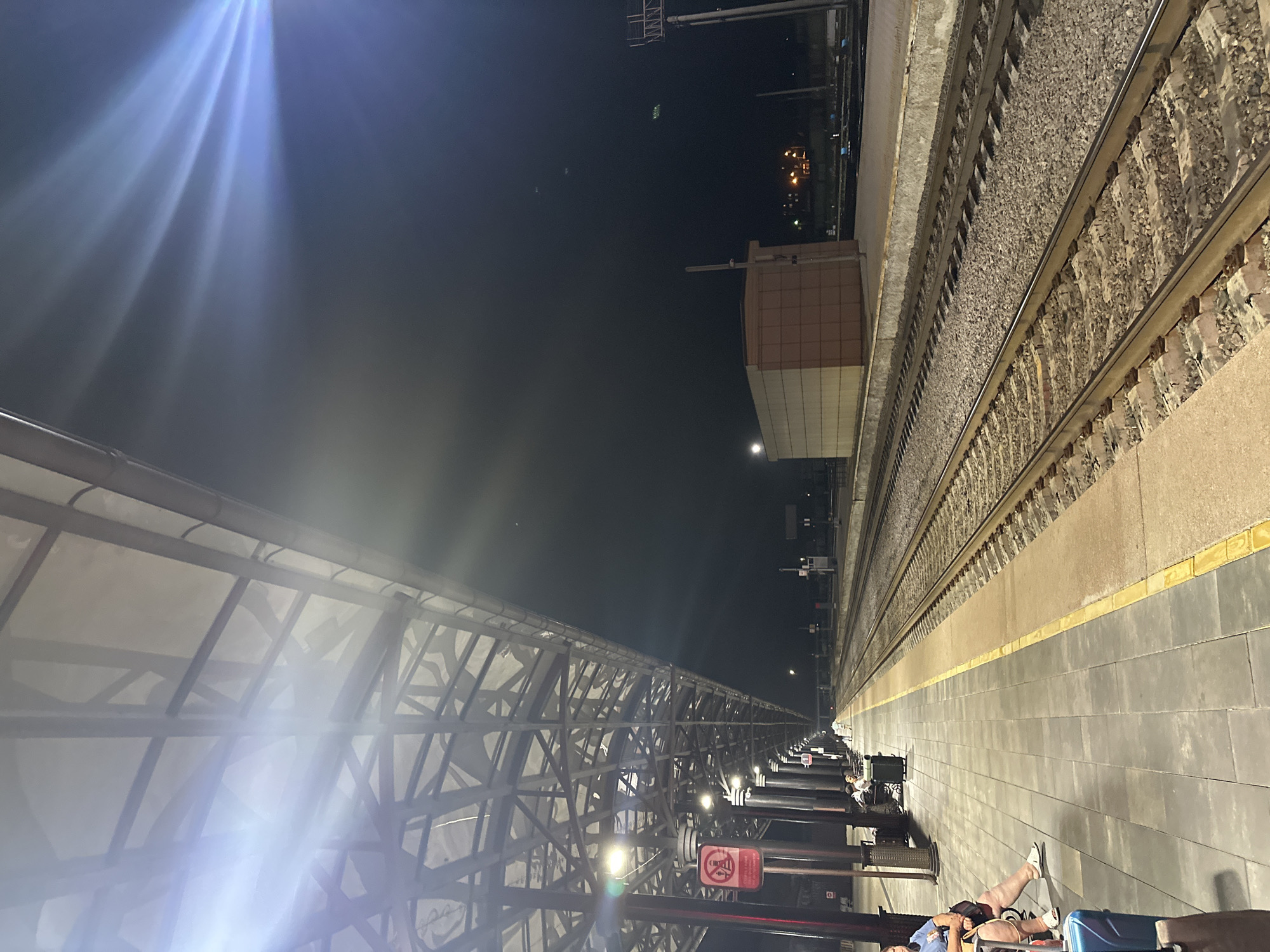 A night time exterior view of a train station platform in Tambov, Russia, shows a covered walkway over a tiled platform with several people sitting on benches. The platform runs alongside multiple sets of train tracks. A brown, rectangular building is visible in the background. Bright lights cast long, lens-flared streaks across the dark sky.