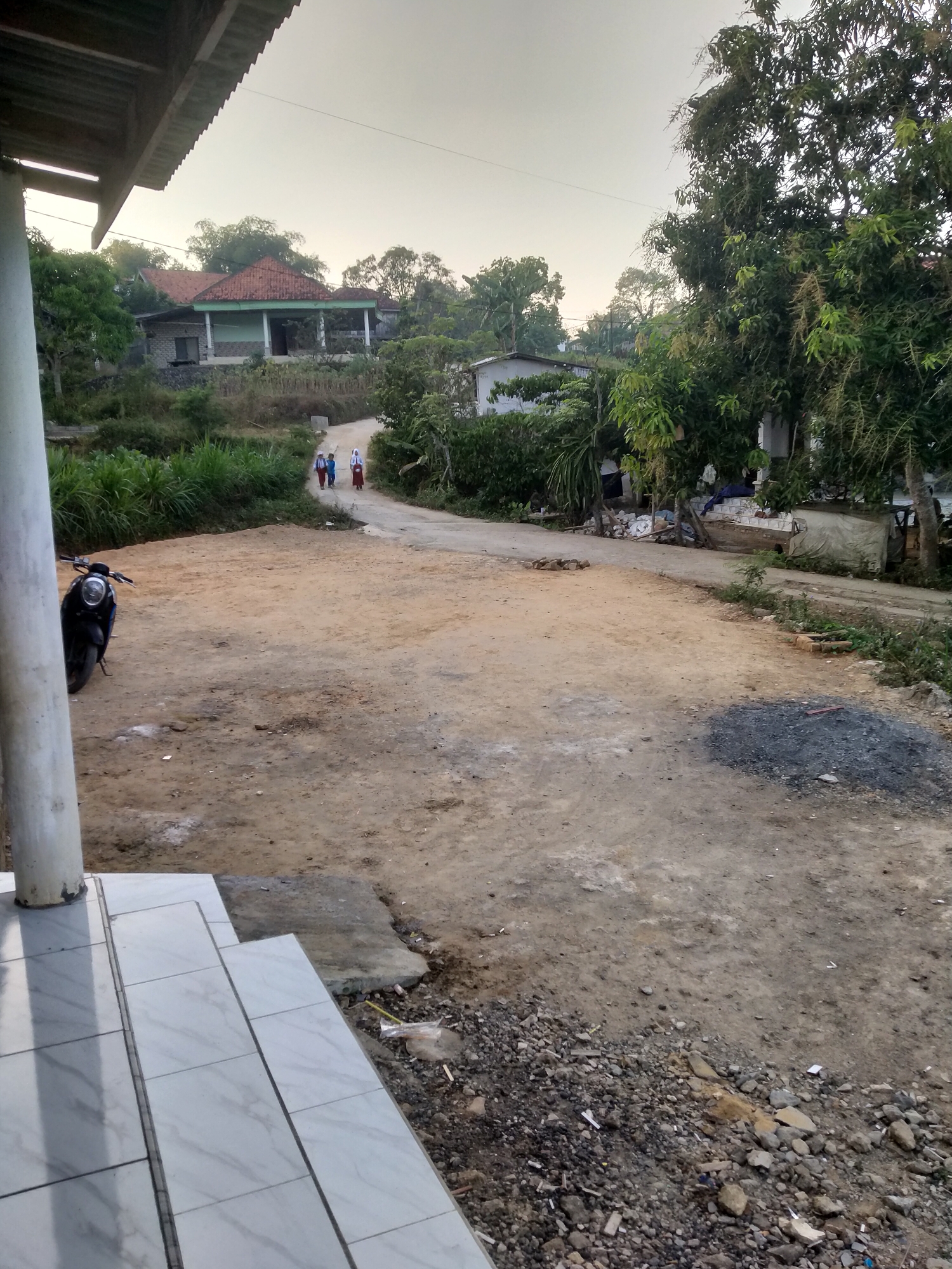 The image depicts a rural road scene in Blaban, Indonesia. A dirt road curves through a landscape with houses, trees, and vegetation. In the foreground, a black motorcycle is parked to the left of the road. Further down the road, three individuals, two appearing to be schoolchildren in uniform, are walking away from the camera. One child is wearing a red and white uniform, and another is in a blue uniform. A third child is dressed in white. The background shows houses with red-tiled roofs and various trees. The sky is overcast, suggesting a cloudy or early morning/late afternoon setting. The overall impression is of a quiet, everyday scene in a rural Indonesian village.