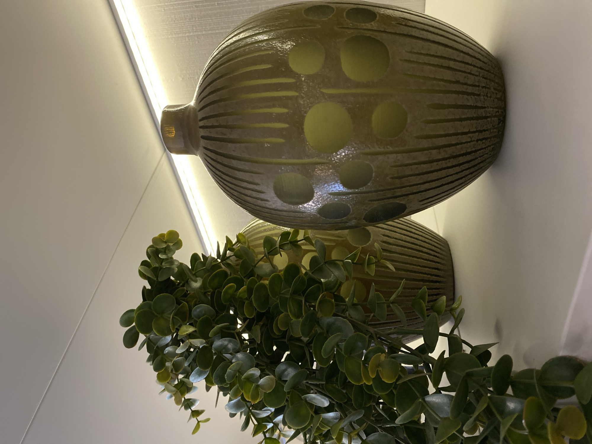 The image features a decorative arrangement on a white surface, possibly a shelf. Two olive-green, ovular vases with circular and horizontal line patterns are stacked, one behind the other. A green plant with small, round leaves cascades down from behind the vases. A bright light source illuminates the scene from above. The image does not provide information about the location or the city of Votkinsk, Russia.
