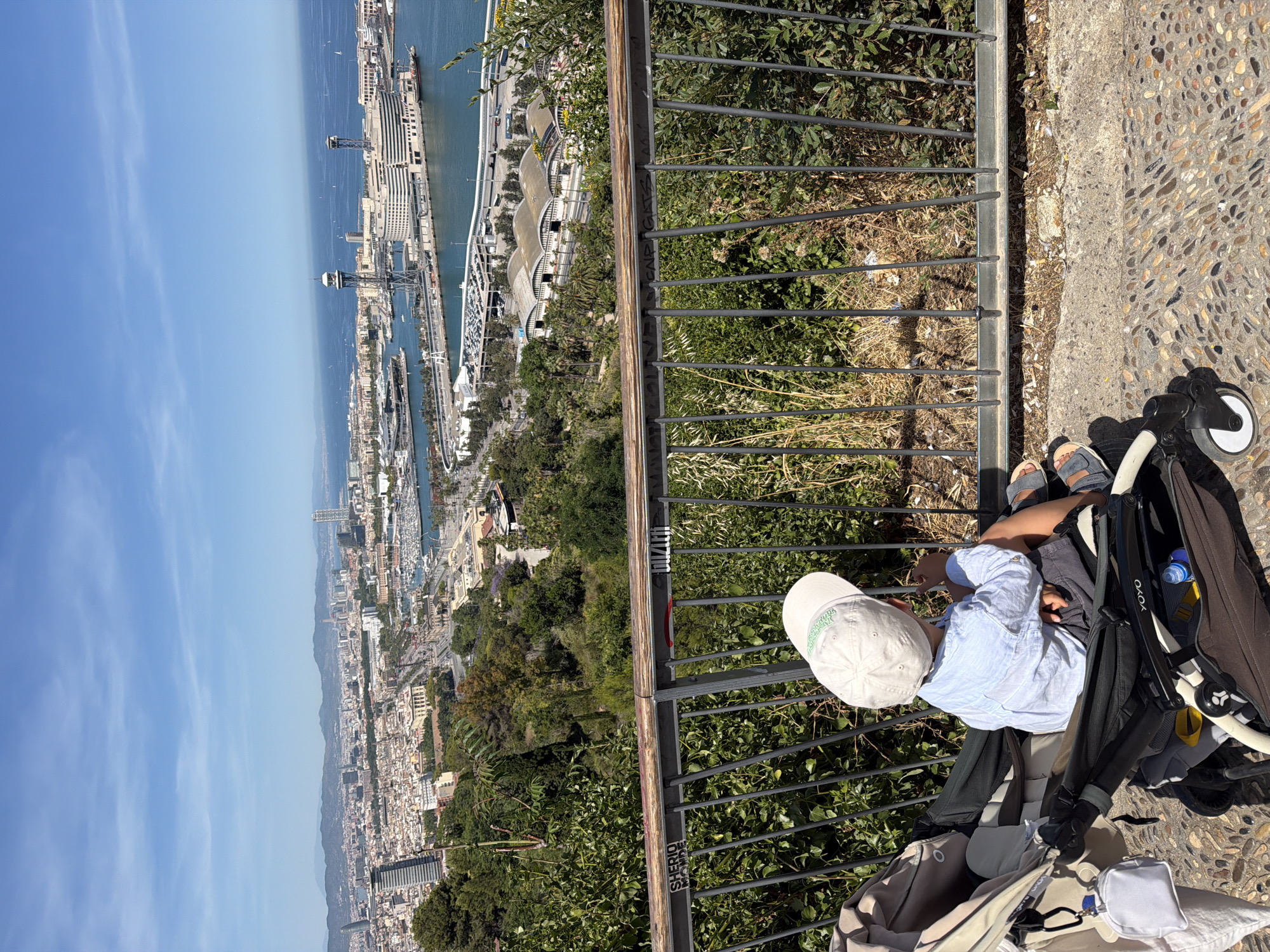 In Barcelona, Spain, from a high vantage point overlooking the city, a young child, wearing a white cap and a light blue long-sleeved shirt, sits in a stroller. The child faces away from the camera, looking out at a panoramic view of the city and the sea. The stroller is dark gray and black, with a white pouch hanging from one of its handles. The child's feet, clad in sandals, rest on the stroller's footrest. A metal railing with graffiti, including the words "SHERIO MANDE" and "DUZIETH", runs horizontally across the foreground, separating the stroller from the lush green foliage below. Beyond the railing, the cityscape stretches out, featuring numerous buildings, a harbor with boats, and the vast expanse of the Mediterranean Sea under a clear blue sky. Mountains are visible in the distance, framing the horizon. The ground beneath the stroller is paved with small, colorful stones.