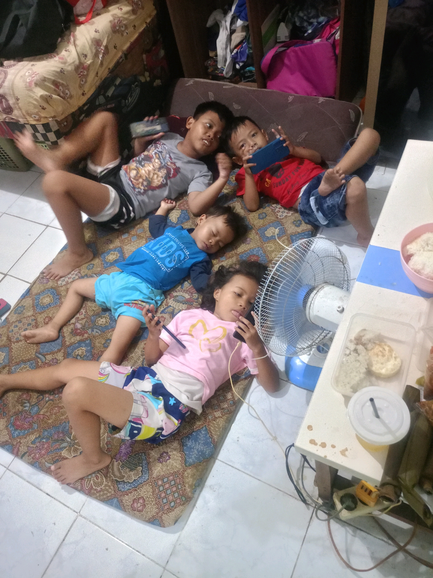 The image depicts four young children lying on a patterned mat on a tiled floor, possibly in a residential setting in Pilangsari, Indonesia. The children are engaged with their mobile phones. Two older boys are positioned at the top of the mat, each holding a phone. A younger boy in a blue shirt and light blue shorts is lying in the middle, with his eyes closed and a phone in his hand. A young girl in a pink shirt and colorful shorts is at the bottom, also looking at her phone.
To the right of the mat, a white table is visible, with a small fan positioned near it. On the table are containers of food, including rice, and a covered drink. Electrical cords are visible on the floor near the table, connecting to an electrical outlet with a yellow adapter. The background shows a cluttered area with furniture and bags, suggesting a living space. The overall impression is of a casual, indoor scene where children are occupied with their devices.