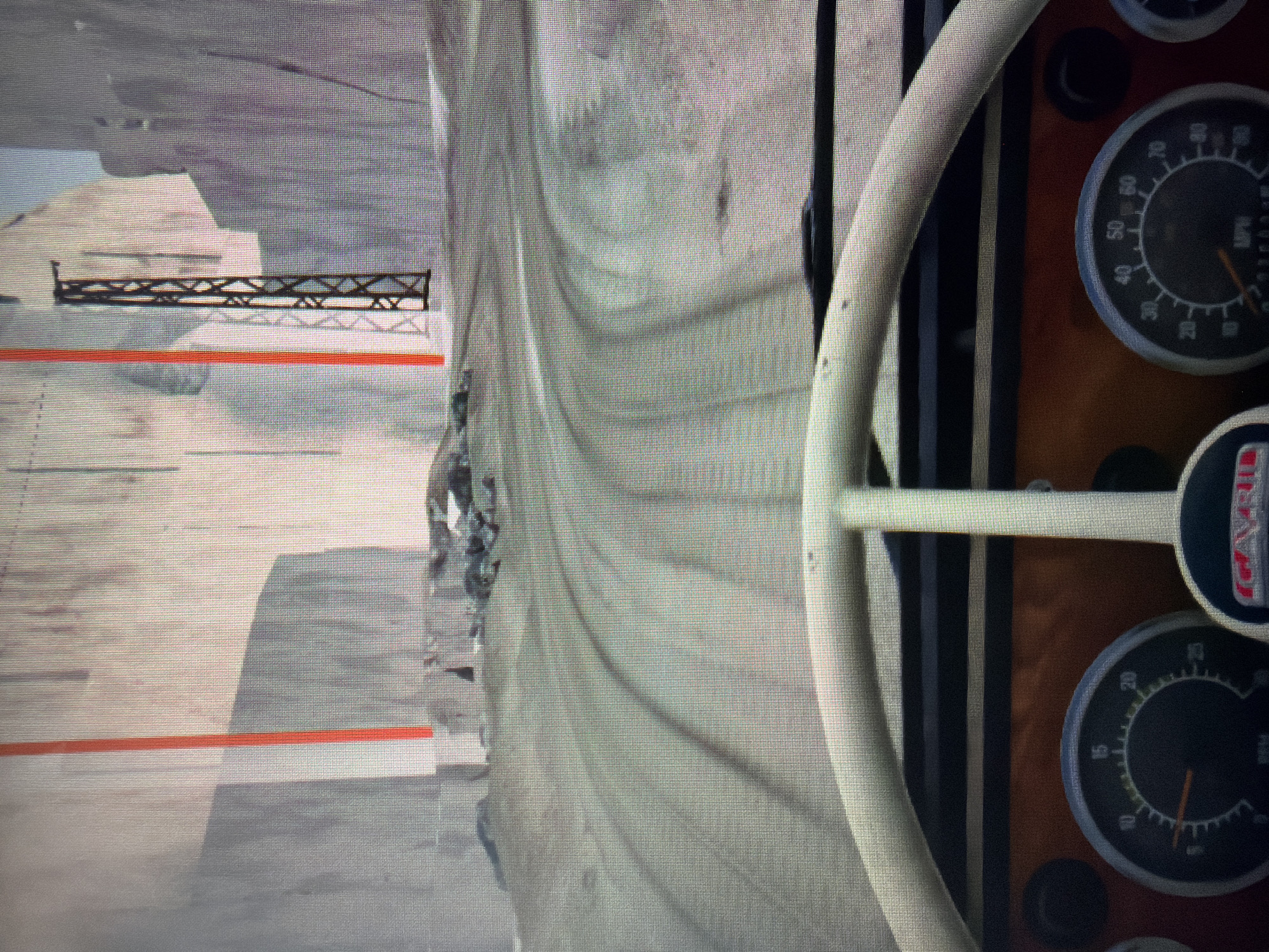 The image shows a view from inside a vehicle, likely a truck, with a steering wheel and dashboard visible. The speedometer reads around 50 mph, and there is another gauge visible below it. Through the windshield, a construction site is visible with piles of dirt or sand and a metal structure, possibly part of a bridge or support system. An orange stripe marks a line on the ground. The location appears to be in an area undergoing construction or demolition, city: Unknown, Unknown. There are no people visible in the image.