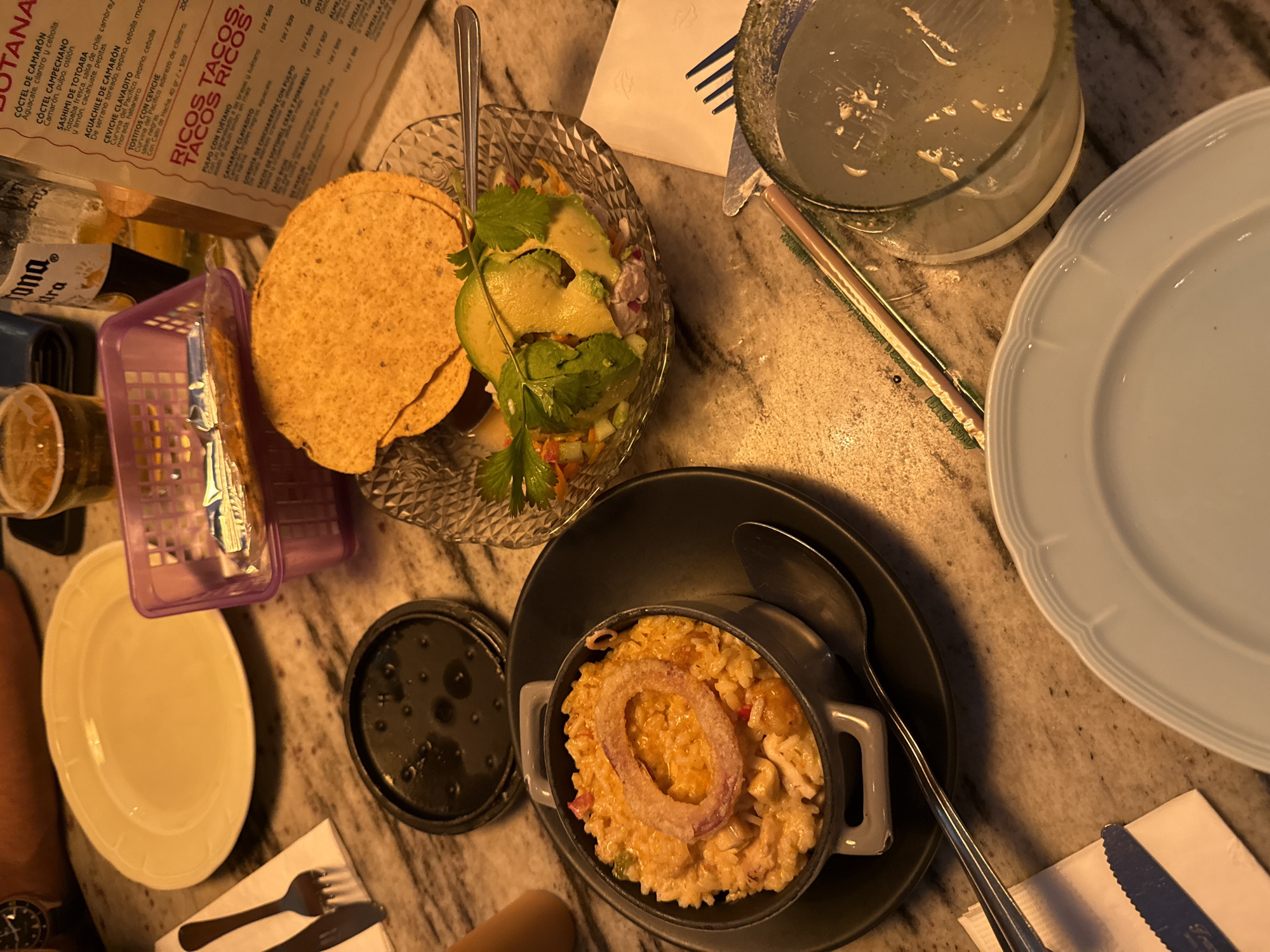 The image shows a table setting with various dishes and beverages. There is a crystal bowl containing what appears to be ceviche with avocado slices, served with tortilla chips. Next to it is a small black casserole dish filled with rice and topped with a fried onion ring, accompanied by a spoon. A glass filled with a liquid, likely a cocktail, is also visible. A light blue plate sits near the glass. The table also has a purple basket, possibly for tortillas, and utensils are placed on a napkin near the plate. A menu card is partially visible, with Spanish text mentioning dishes like "COCTEL DE CAMARÓN," "CEVICHE CLAVADITO," and "RICOS TACOS, TACOS RICOS." The image also shows parts of a person's arm and wrist with a watch.
Based on the menu items and language, the location is likely a restaurant serving Mexican cuisine, specifically in San Pedro Garza García, Mexico.