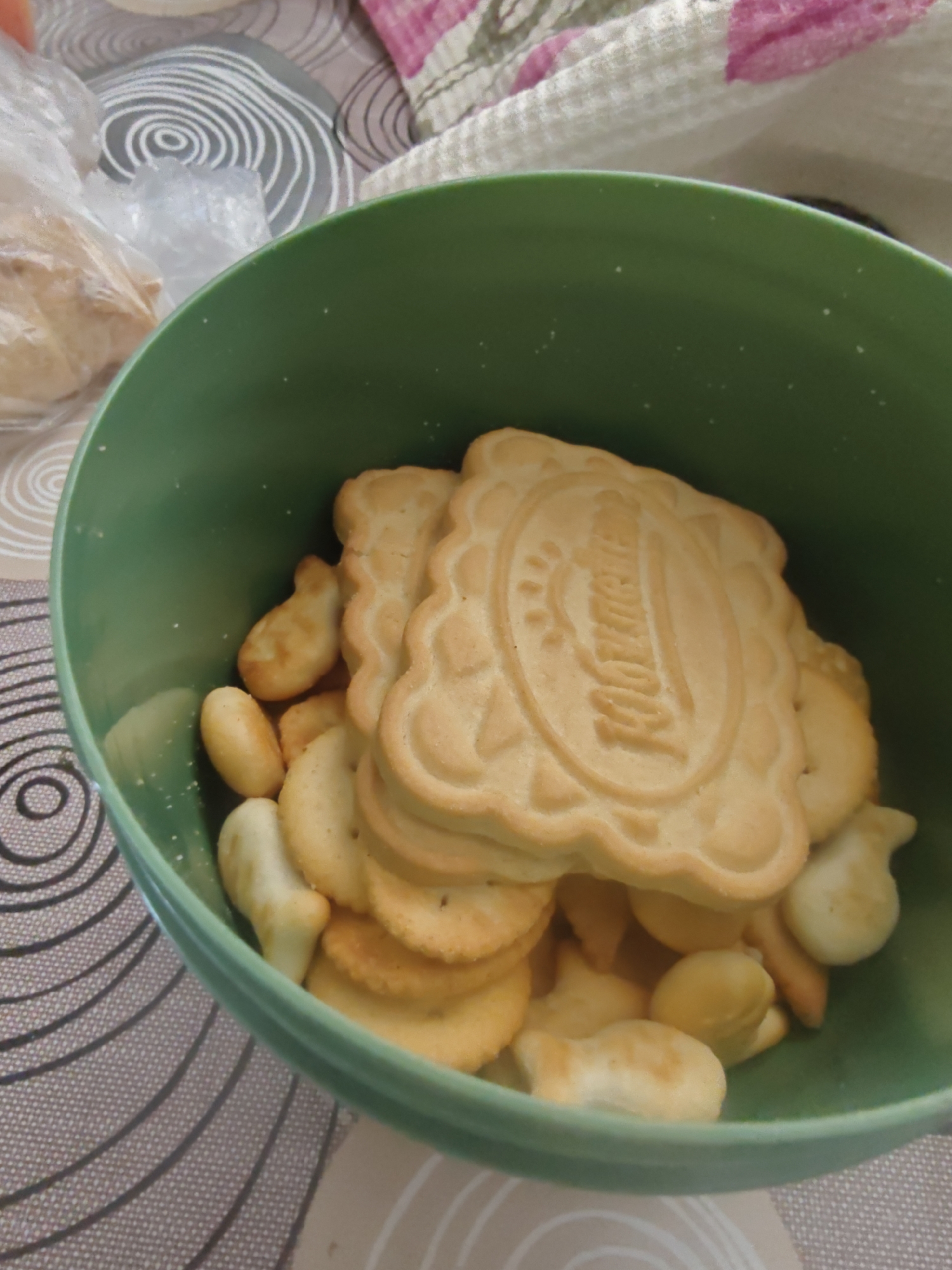 A close-up shot shows a green bowl filled with crackers, including gold fish crackers and larger, rectangular crackers. The crackers are golden brown and appear to be lightly salted. The bowl is on a patterned surface, and a portion of a plastic bag containing some bread is visible to the left. A patterned cloth is also in the background. The image appears to be taken indoors. There is no visible information within the image to suggest a specific location in Krasnodar, Russia.