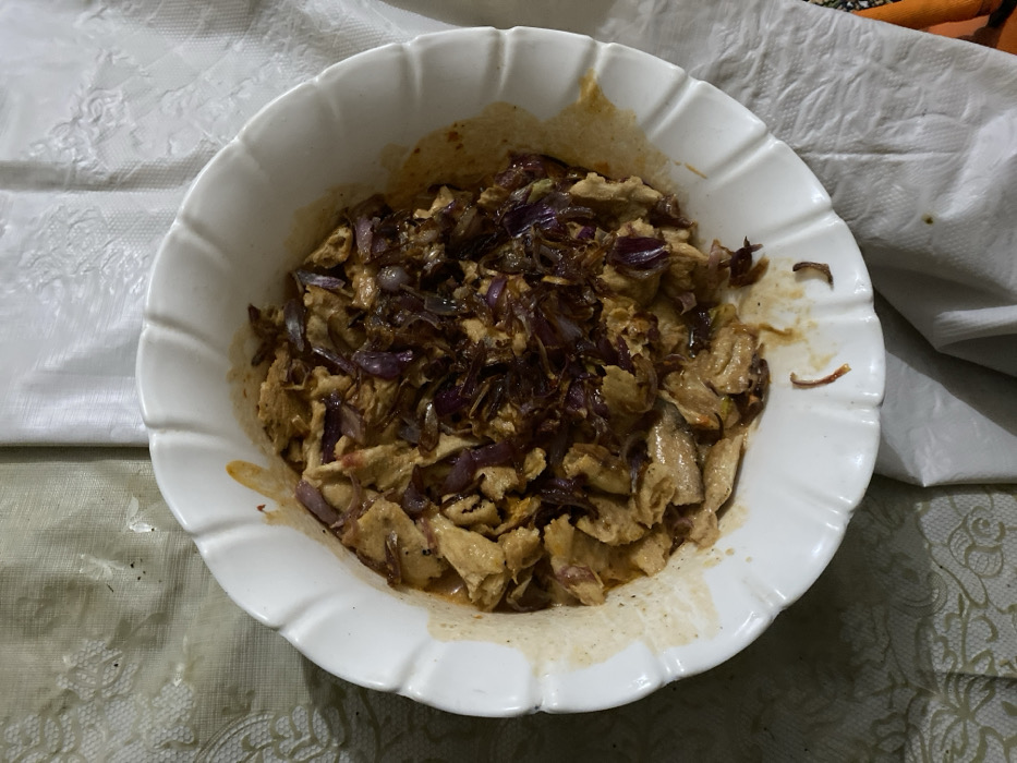 A white, fluted bowl holds a mixture of sliced cooked chicken and sautéed red onions. The chicken appears to be lightly seasoned or sauced, with a slightly golden hue. The red onions are caramelized, adding a rich color contrast and flavor element. The bowl sits on a tablecloth with a subtle floral pattern, with a white, wrinkled fabric or napkin nearby. Part of an orange object is visible in the upper right corner.