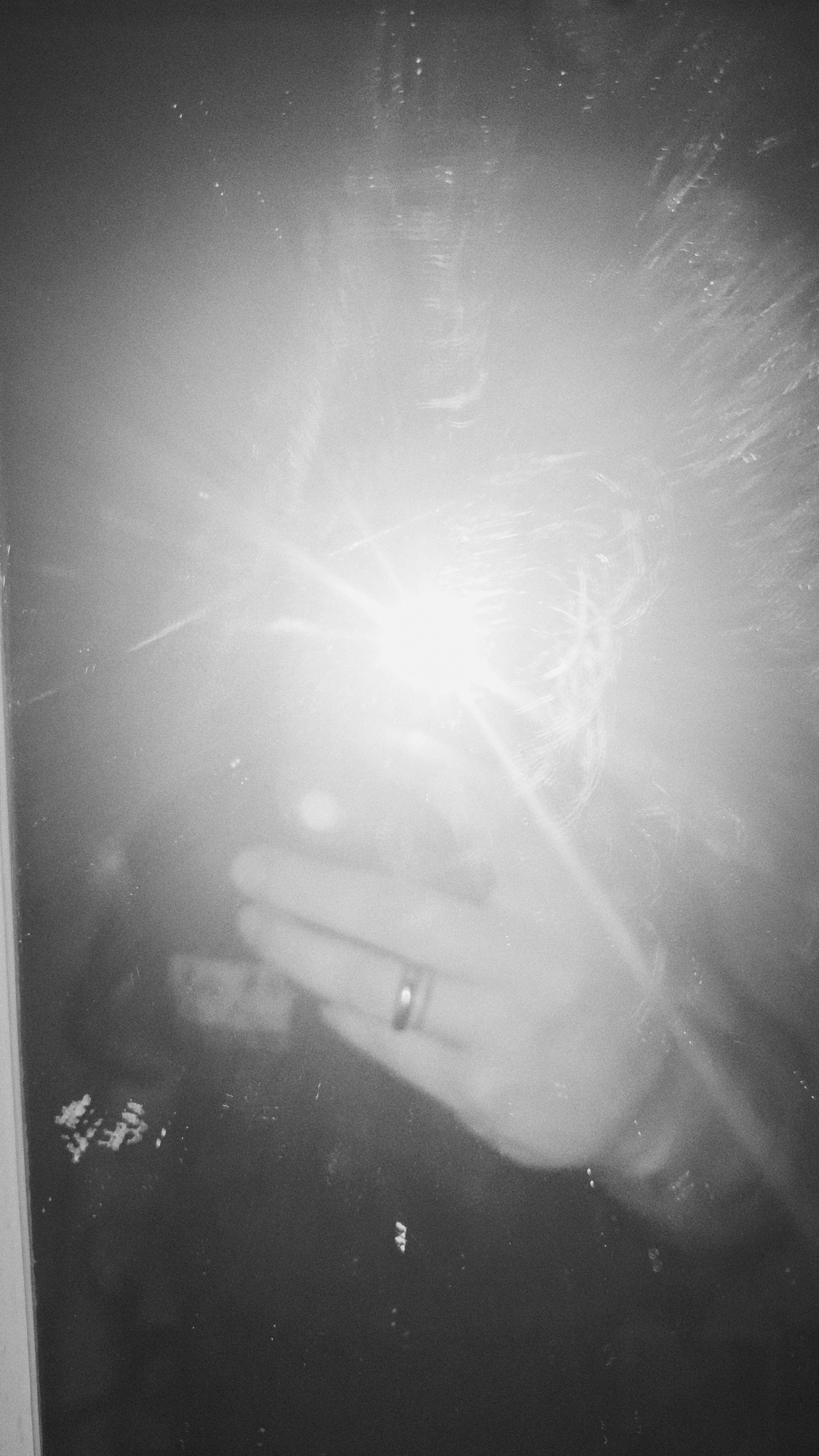 The image is a black and white photograph of a hand holding a light source, possibly a smartphone or flashlight, which causes a bright glare obscuring much of the scene. A ring is visible on one of the fingers of the hand. The background is dark and indistinct, with some grainy texture. It is difficult to determine the location in Nowy Tomyśl, Poland or other specific details due to the strong light and lack of discernible features.