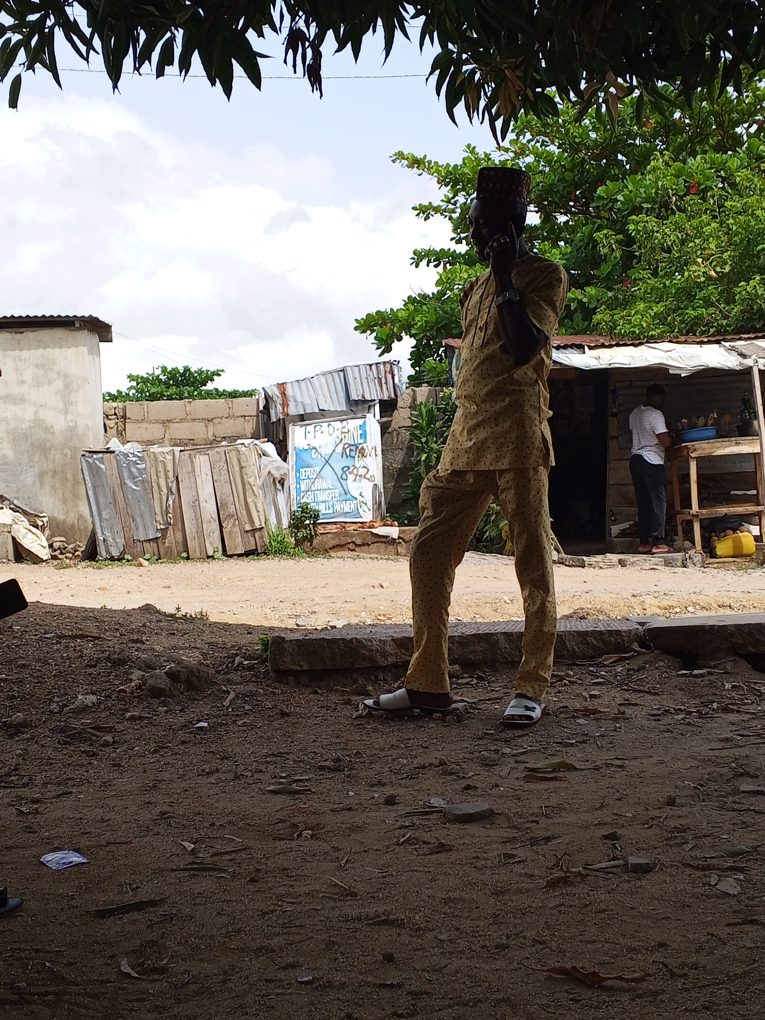 The image shows a man standing on a dirt road in what appears to be a rural area. He is wearing a traditional Nigerian outfit and holding a phone to his ear. Behind him, there are several buildings, including a small shop where another man is standing behind a counter. The area is littered with debris and appears to be in a state of disrepair. The location is Zango-ndaji, Nigeria.