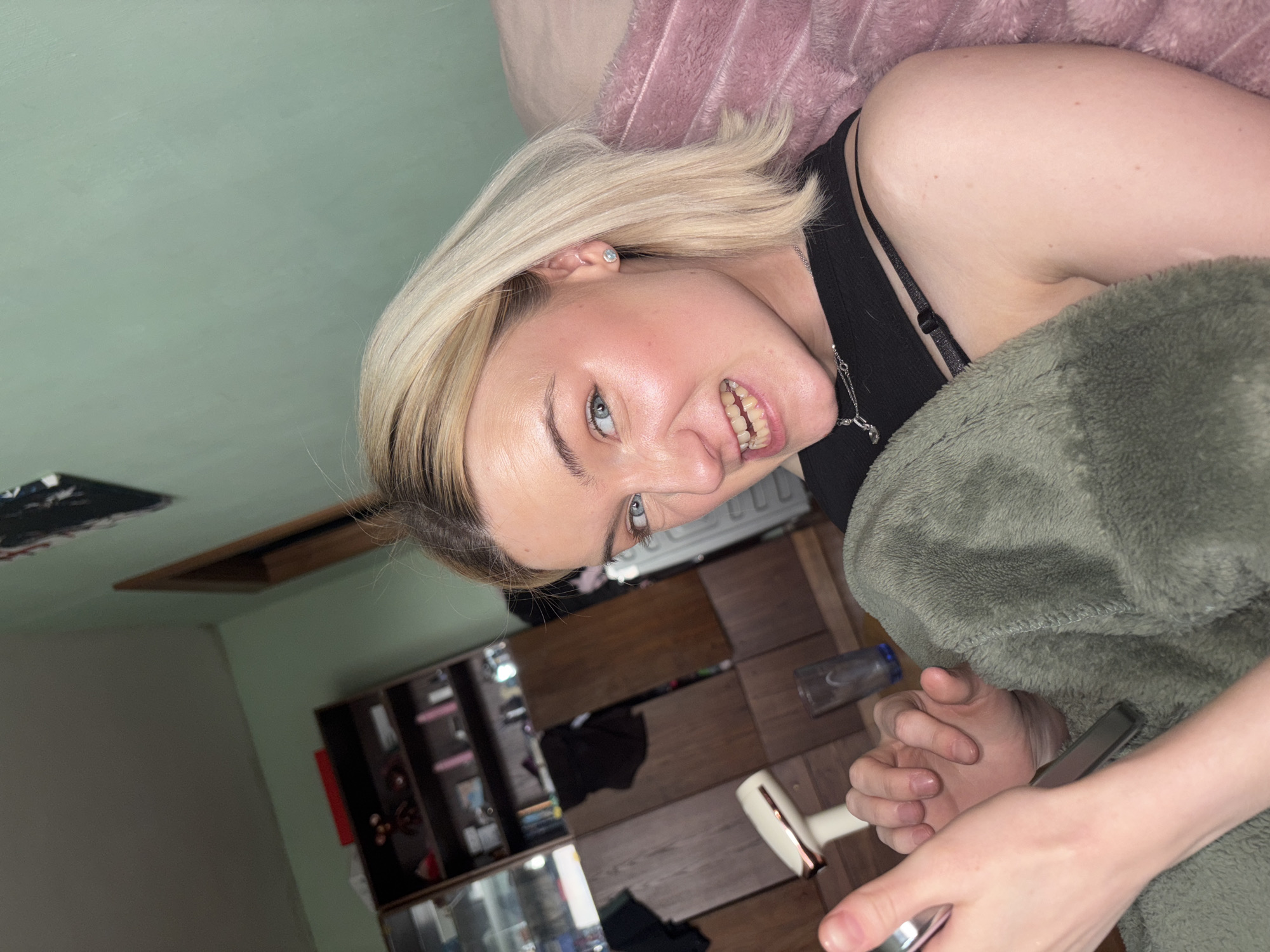 A young person, likely female, with blonde hair and blue eyes, is shown in an indoor setting, possibly a bedroom in Vsevolozhsk, Russia. The individual is reclining or lying down, looking up at the camera with a wide, playful smile that reveals their teeth. They are wearing a black tank top or camisole, with a silver-colored necklace and a small stud earring in their left ear.
Their body is partially covered by a fluffy, olive-green blanket, with a pink, similarly textured blanket or pillow behind their head. Their hands are visible in the foreground, holding a silver-colored smartphone or small device.
The background features light green walls. An opening in the wall or ceiling, possibly an attic access or ventilation, is visible on the left. In the mid-ground, wooden furniture, resembling a chest of drawers or shelving unit, can be seen, holding various items. A clear plastic bottle and a white, possibly electronic, object (like a fan) are on the floor. The lighting suggests an indoor setting, and the person's expression conveys amusement or lightheartedness. No discernible text is visible in the image.