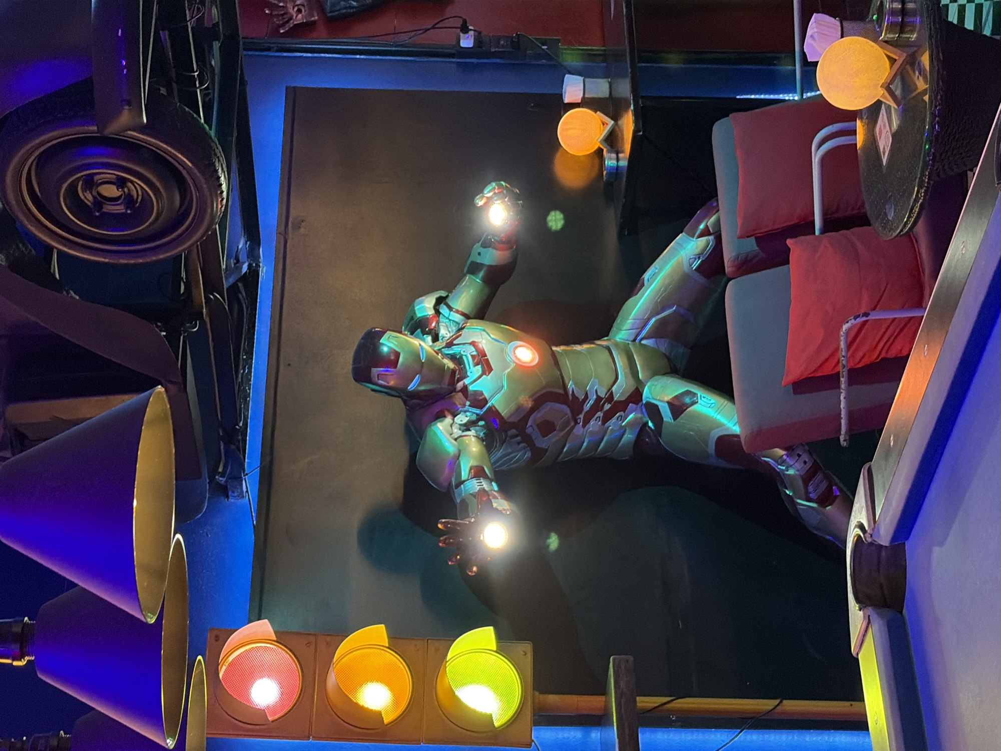 In Baan Lamai, Thailand, an Iron Man statue is displayed in what appears to be a themed entertainment venue. The statue is positioned in a dynamic, action-ready stance, with its hands glowing, suggesting the use of its repulsor rays. The environment is dimly lit, with vibrant blue and purple lighting accentuating the scene. Hanging above are large, conical light fixtures and a car tire, possibly part of the decor. To the left, a traffic light display with illuminated red, yellow, and green bulbs adds to the quirky atmosphere. In the foreground, elements of a pool table are visible, hinting at recreational activities. The background features plush red chairs and a small table with a glowing moon-shaped lamp, creating a lounge-like setting.