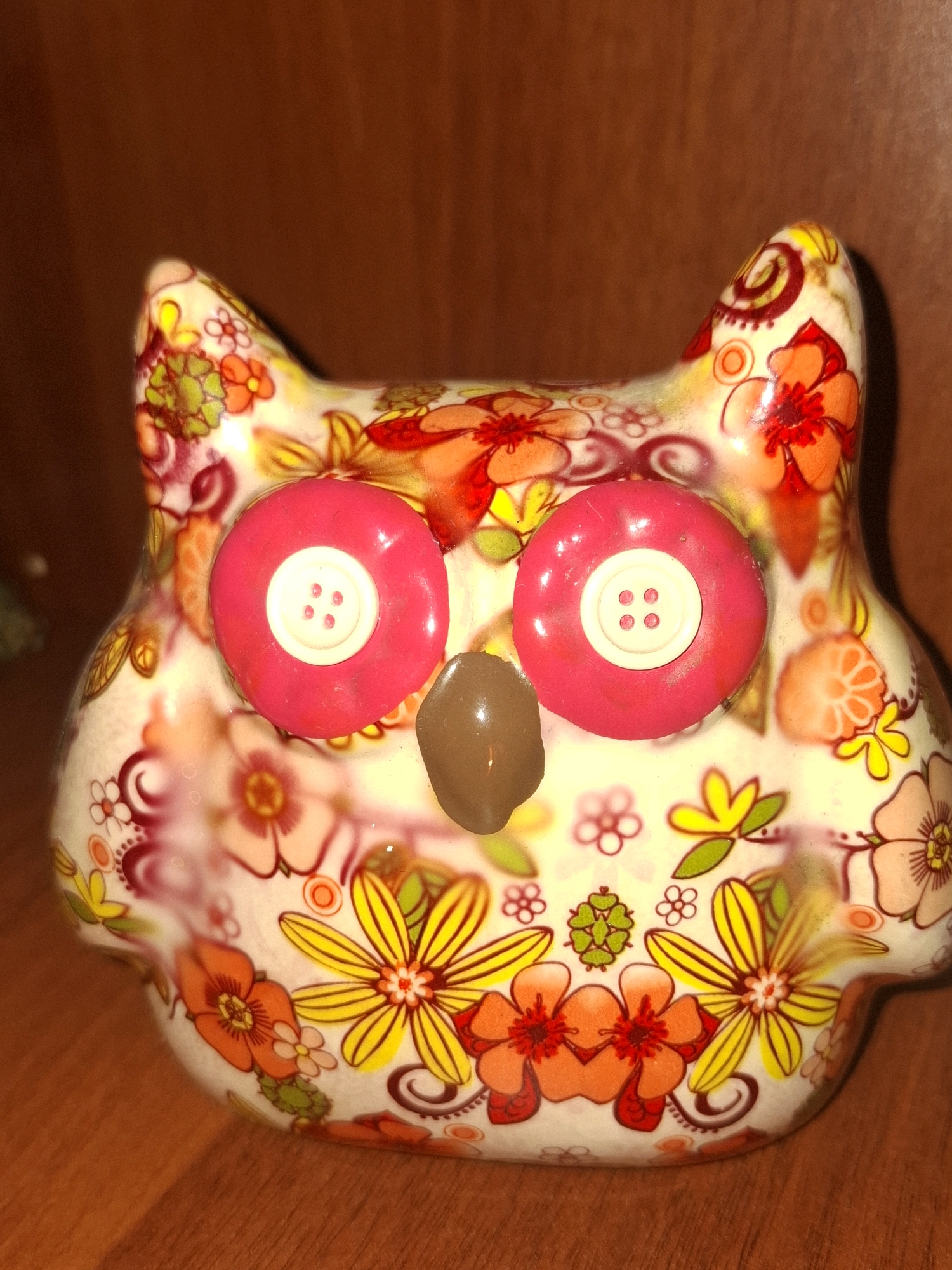 A close-up, head-on, vertical shot displays a decorative ceramic owl figurine with a floral pattern. The owl has large, pink, button-like eyes with white centers and four holes each. A small, brown, oval beak is centered between the eyes. The figurine's surface is covered in a vibrant, colorful pattern of various flowers, leaves, and swirls in shades of orange, yellow, red, green, and pink against a cream-colored background. The owl is positioned on a wooden surface, likely a table or shelf. The background is a blurred, warm-toned brown, suggestive of a wooden wall or furniture. The lighting is warm and direct, highlighting the glossy finish of the figurine and casting subtle shadows. The image does not contain any information that would allow for the identification of a specific location within the city of Almaty, Kazakhstan.