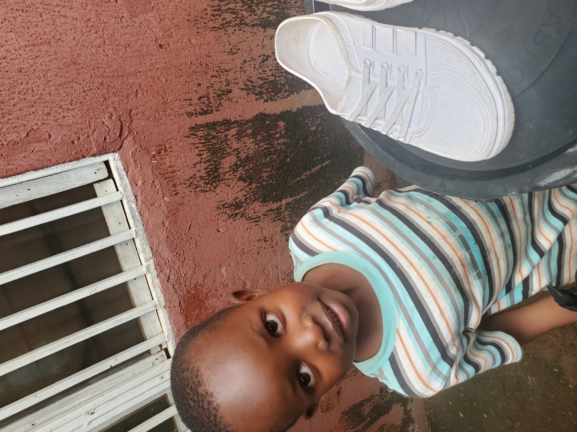 A young child, likely a boy, is the primary subject of this medium shot, captured from a slightly elevated, angled perspective. He is wearing a horizontally striped t-shirt in shades of blue, white, orange, and gray. The child is looking directly at the camera with a slight smile, his dark eyes conveying a sense of openness and innocence. His dark hair is cut short.
To the right of the child, a white shoe is visible, resting on a large, dark circular object, possibly a bucket or bin. The shoe appears to be made of a white synthetic material, possibly plastic or rubber, with laces. The background consists of a rough, reddish-brown textured surface, which appears to be wet in places, suggesting recent rain. To the left of the child, there is a white slatted structure, resembling a window grate or a fence.
The scene appears to be outdoors, given the wet ground and the reddish-brown wall, which could be an exterior wall. The lighting suggests it is daytime. The wet surfaces hint at recent rain, implying a humid or tropical climate, consistent with the provided location context of Jos, Nigeria. There is no visible text in the image. The overall impression is of a candid moment with a child in an outdoor, possibly domestic, setting.