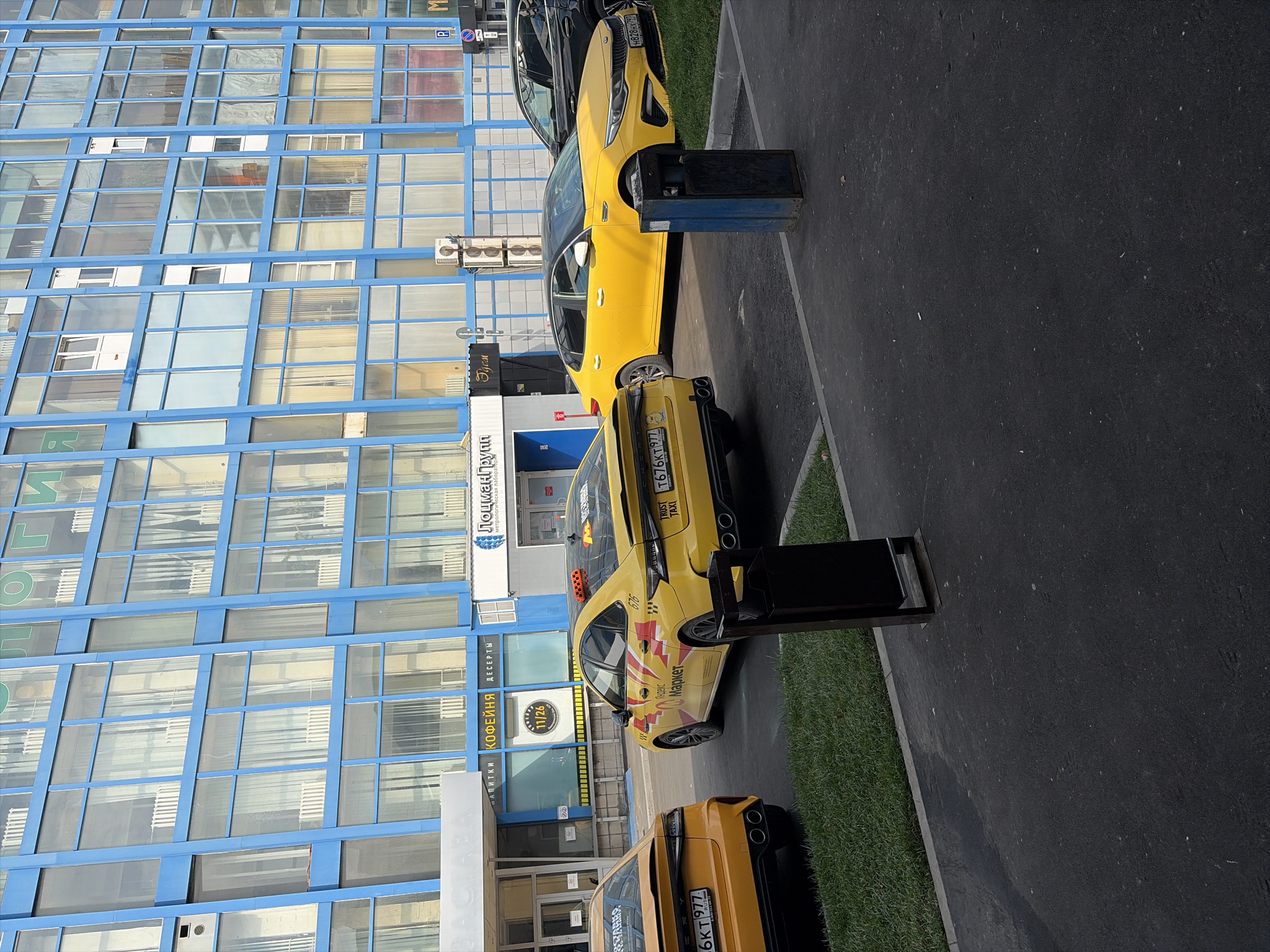 The image depicts a street scene in Moscow, Russia, featuring several yellow taxi cars parked in front of a modern blue building with numerous windows. The building appears to be an office or commercial complex. The taxi cars are modern sedans, with one prominently displaying "Яндекс Такси" (Yandex Taxi) branding. One car has the license plate "T676KT 77 RUS", another "H828HY 77 RUS", and a third "6KT 977". There are also trash receptacles visible on the asphalt. The weather appears to be clear and sunny. No people are clearly visible or interacting with the vehicles or environment.