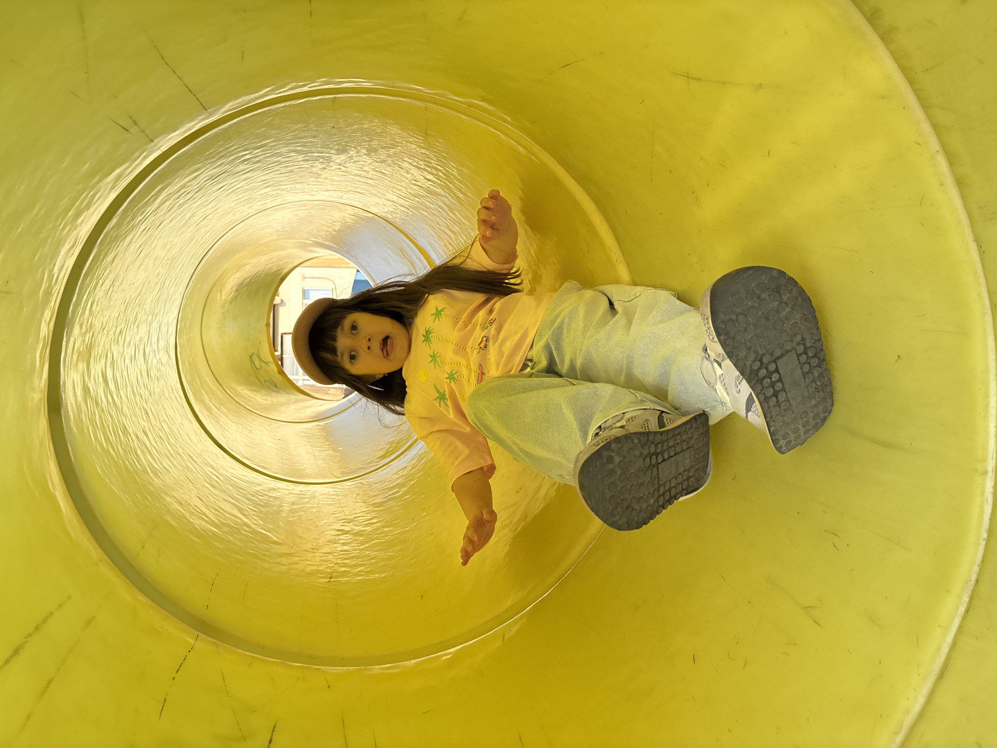 The image shows a young girl with dark hair, wearing a yellow t-shirt with palm tree graphics and a light-colored baseball cap, sliding down a bright yellow tube slide. She is wearing light-colored pants and dark-soled sneakers. Her mouth is open in an expression of surprise or excitement. The interior of the slide is brightly lit and appears to be made of plastic. The background, visible at the end of the tube, suggests an outdoor setting, possibly a playground, with glimpses of buildings. The image was taken in Chaikovsky, Russia.