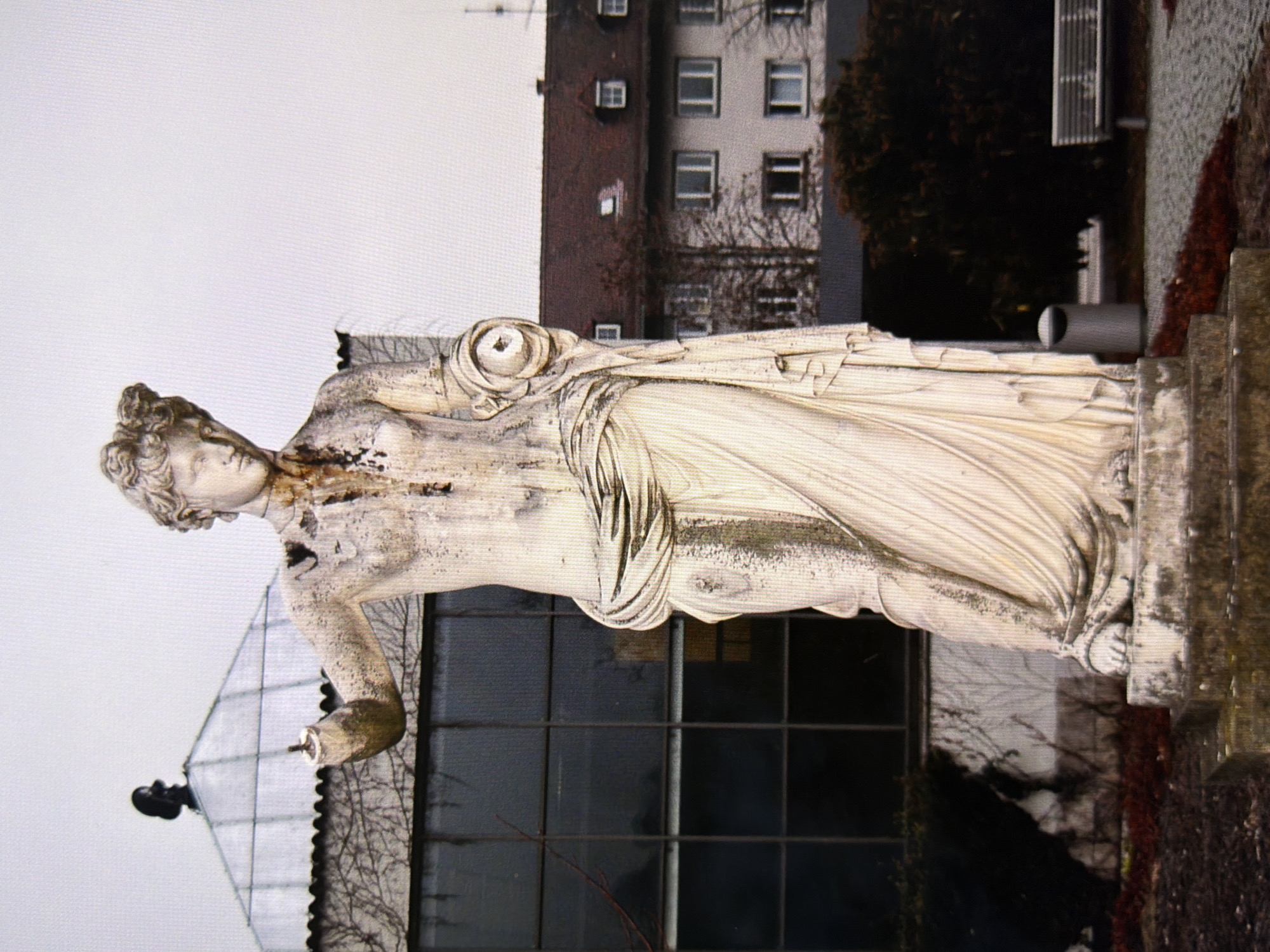 An outdoor, full-shot photograph shows a weathered, classical-style stone statue of a woman in a draped gown. The statue is partially damaged, with missing portions of its arms and some discoloration and moss growth. In the background, there is a large, dark-paned glass building with a peaked roof, partially obscured by bare tree branches. Further in the background, a multi-story building with a red-tiled roof and numerous windows is visible. The ground in front of the statue is covered with dark red mulch, and to the right, a modern park bench and a trash receptacle are present. The overall scene is set in an urban environment, likely a park or public space. The image is of poor quality with visible pixelation. The location of this statue is not specified in the image or its metadata.