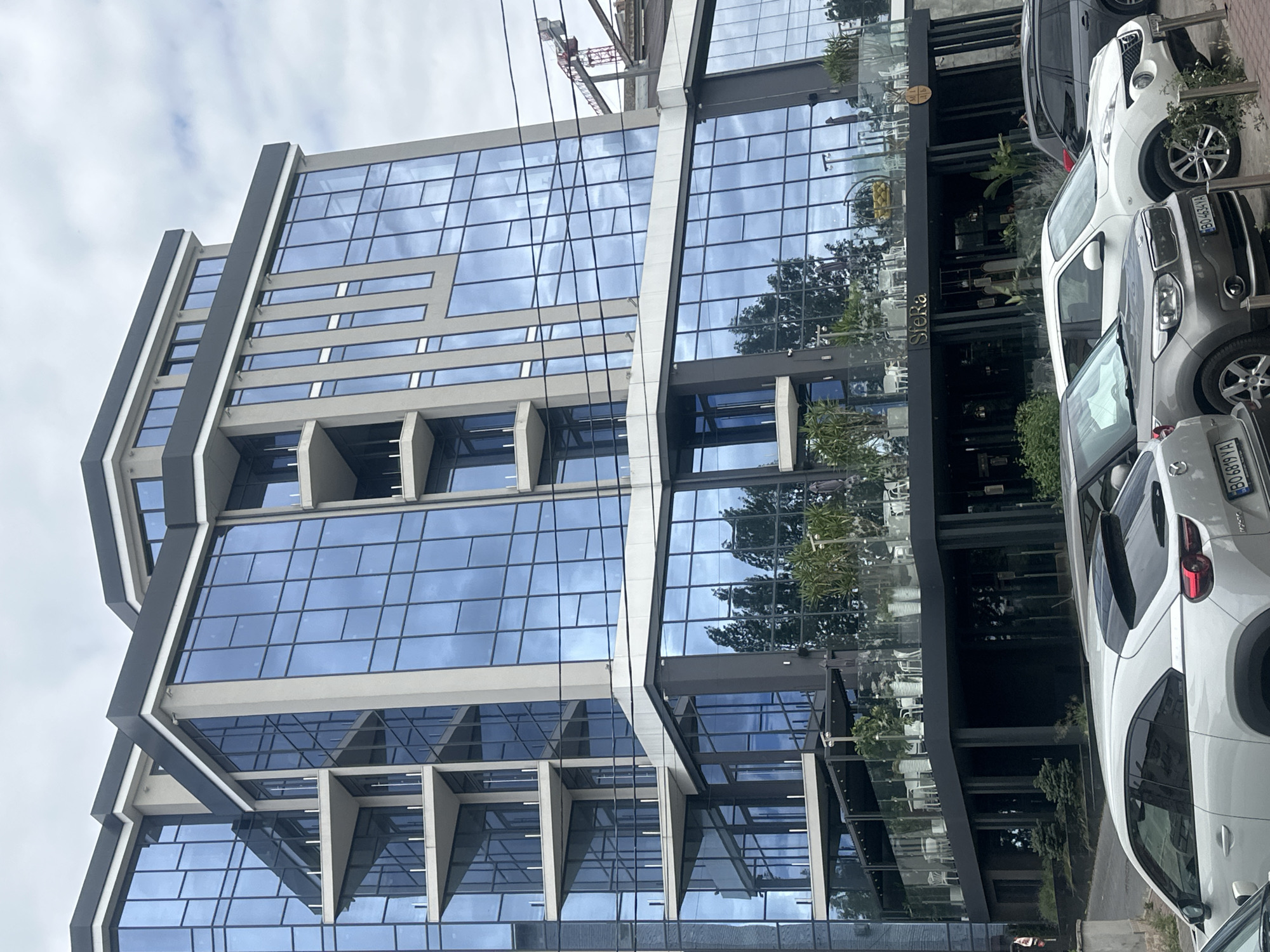 The image shows a modern, multi-story building with a glass facade in Ternopil, Ukraine. The building has a sleek, contemporary design with large, reflective blue windows that mirror the cloudy sky and surrounding trees. Several balconies with glass railings are visible on different levels, adding to the building's architectural style. The ground floor appears to house a commercial establishment, possibly a restaurant or cafe, with outdoor seating and decorative plants. A sign that reads "Sfera" is visible on the building. Several cars are parked in front of the building, suggesting an urban environment. In the background, a construction crane is visible, indicating ongoing development in the area. The overall scene depicts a well-maintained urban setting with a focus on modern architecture.