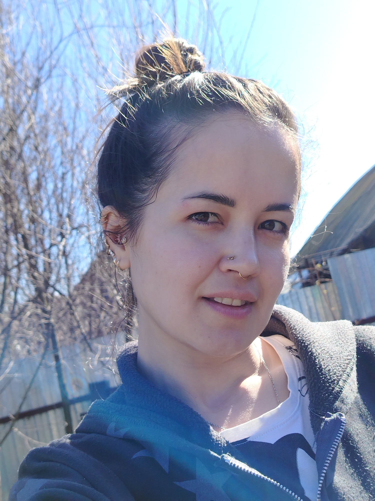 A selfie of a woman with dark hair pulled up into a bun on the top of her head. She has light skin, dark eyes, and multiple ear piercings. She also has a nose piercing. She is wearing a white t-shirt with a black design, and a gray hoodie. The background is blurry, but includes bare tree branches, a gray fence, and a corrugated metal building. The location is possibly Гожан, Russia.