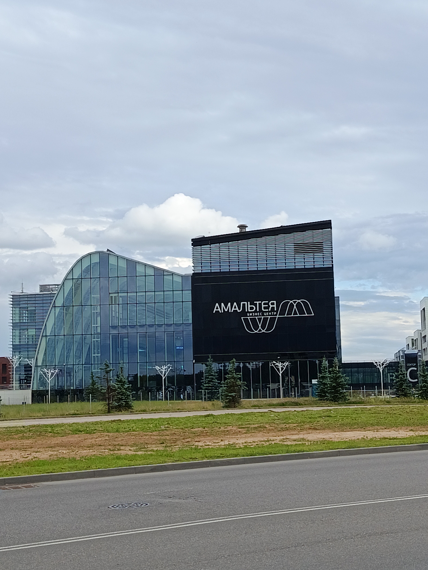 This is a photograph of a modern business center on a cloudy day. The building on the right is predominantly black and features a large white logo and text that reads "АМАЛЬТЕЯ БИЗНЕС ЦЕНТР". To the left of this is a curved building with a glass facade, reflecting the sky and surrounding greenery. Several evergreen trees are planted in front of the buildings, and a strip of grass and a road are visible in the foreground. The sky is overcast with a mix of light and dark clouds. The overall impression is one of a commercial or office environment on a typical day. There are no people visible in the image. The scene appears to be in an urban or suburban setting.