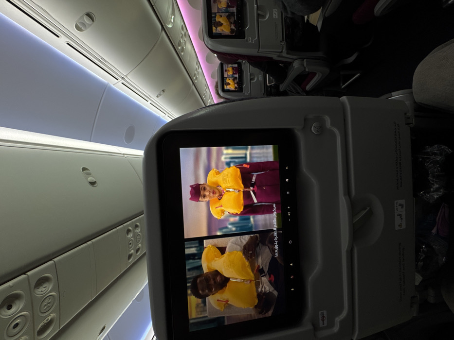 The scene depicts the interior of a commercial passenger aircraft, likely on a flight associated with Zanzibar, Tanzania. The image is captured from a passenger's viewpoint, looking forward towards the seat in front and down the cabin aisle.
In the immediate foreground, the back of a standard aircraft seat is visible, featuring a rectangular in-flight entertainment (IFE) screen. This screen displays a split-image safety video. On the left side of the screen, a Black male, identified as actor Kevin Hart, is shown from the chest up, wearing an inflated yellow life vest. On the right side, a light-skinned female flight attendant, dressed in a maroon uniform with a matching cap, is also depicted from the chest up, wearing an inflated yellow life vest. Arabic text is present below the images on the screen. Below the screen, the folded tray table mechanism is visible, along with text instructions in English and Arabic. The legs of the person taking the photograph are partially visible at the bottom of the frame, dressed in light-colored trousers, with personal items stored on the floor under the seat in front.
Further into the cabin, multiple rows of passenger seats extend towards the background. Each visible seatback also features an active IFE screen displaying content consistent with the safety video. Passengers are partially visible in some seats. The overhead cabin features white panels, integrated lighting fixtures, and passenger service units. White ambient lighting illuminates the main cabin, complemented by strips of magenta-colored accent lighting running along the ceiling on the right side of the cabin. The overall environment indicates an active flight segment where passengers are viewing safety instructions.