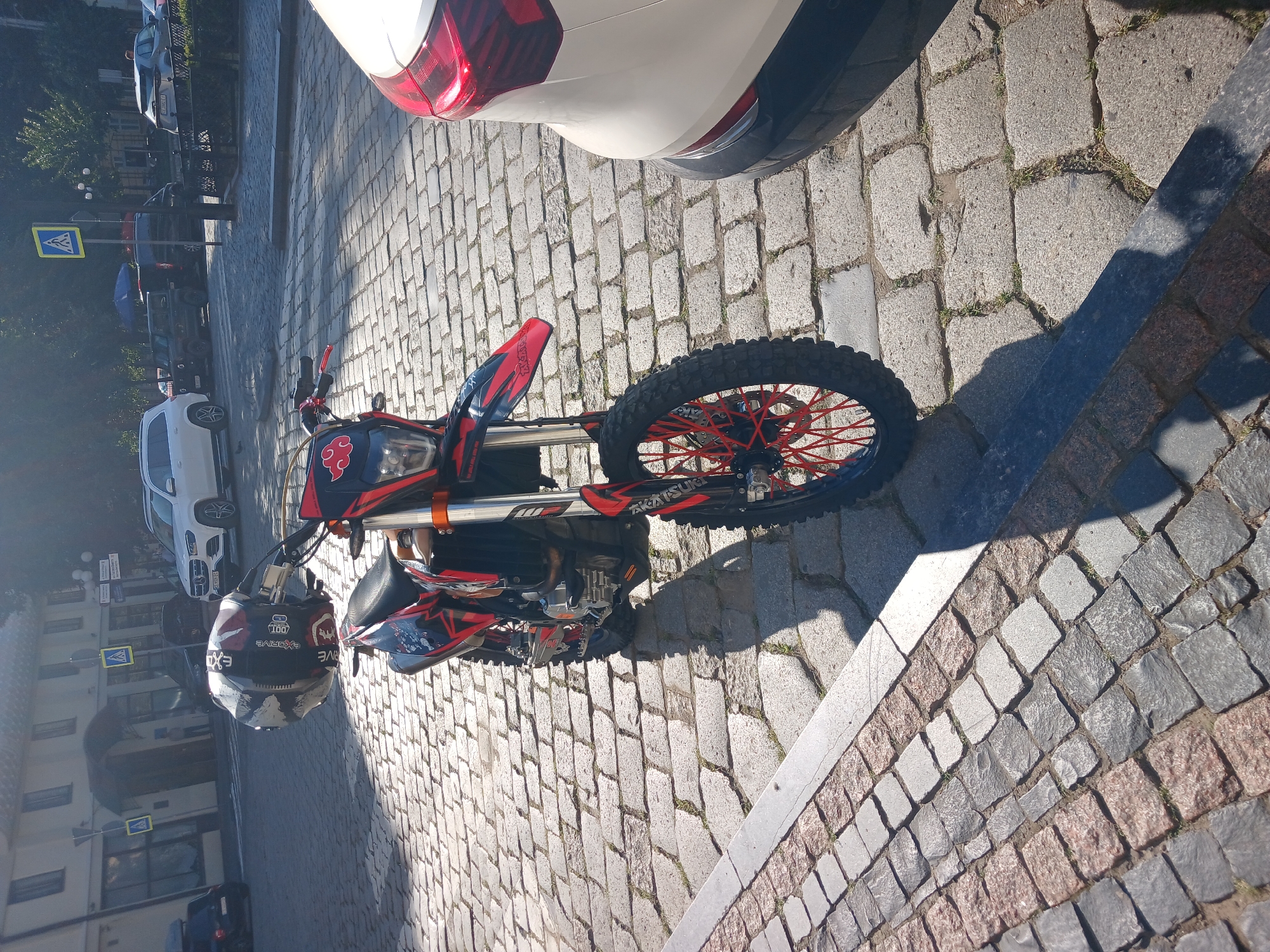 A dirt bike is parked on a cobblestone street in Chernivtsi, Ukraine, casting a shadow to the right. The motorcycle has red and black graphics and a helmet rests on its handlebars. A white SUV is parked behind the motorcycle, and other vehicles and buildings are visible in the background. The scene appears to be a sunny day in a city square.