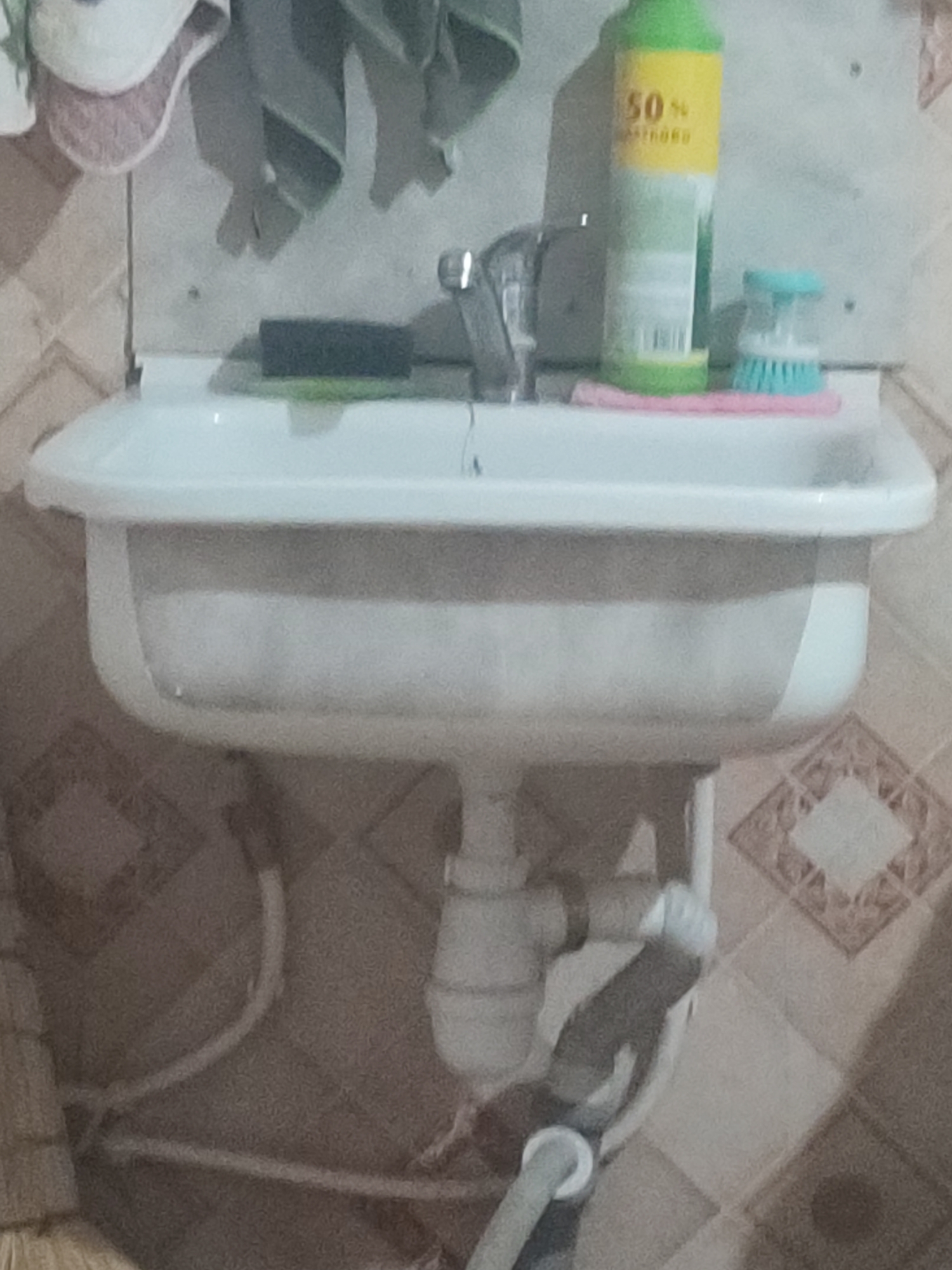 A slightly blurry, medium-angle shot captures a white porcelain sink with plumbing visible underneath. To the right of the faucet, on the sink's edge, sits a bottle with a green label and yellow text that appears to read "50%". Next to the bottle is a small blue and white scrub brush. The sink and its accessories are set against a tiled wall with a diamond pattern. The scene suggests a domestic bathroom or utility area. There are no people visible. The location is the city of Chernivtsi, Ukraine.
