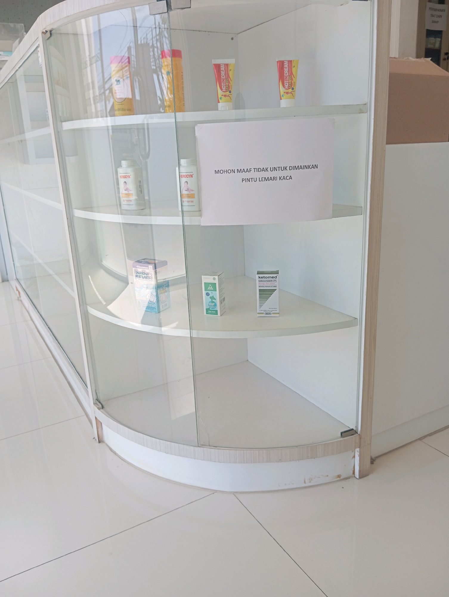 This is a photograph of a glass-fronted display cabinet, likely in a pharmacy or convenience store, in Sumampir, Indonesia. The cabinet is curved and has multiple shelves. On the top shelf, there are three tubes of cream, two of which appear to be "HOTIN CREAM" with yellow and red labels, and another with a yellow label. Below this, on the second shelf, are two white bottles with red caps, each labeled "HERO-CYN" with an image of a baby. Also on this shelf is a white paper sign with black text that reads "MOHON MAAF TIDAK UNTUK DIMAINKAN PINTU LEMARI KACA" which translates to "SORRY, DO NOT PLAY WITH THE GLASS CABINET DOOR." On the third shelf, there are two small boxes. One is blue and white, labeled "Bye Bye Fever." The other is a smaller white box with green accents, labeled "ketomed ketoconazole 2%". The cabinet is situated in a brightly lit indoor space with white tiled flooring. The background shows reflections in the glass and a portion of a wall with some shelving and boxes.