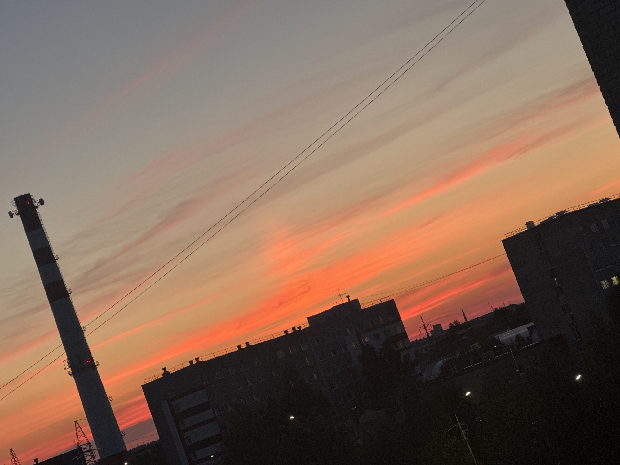 A drone captures a low-angle shot of a vibrant sunset over Surgut, Russia. The sky is a gradient of pale gray at the top, transitioning to soft pink and fiery orange as it descends towards the horizon. Wispy clouds are illuminated by the setting sun, creating streaks of color across the sky.
Dominating the left side of the frame is a tall, industrial smokestack, its upper section painted in alternating red and white bands. It is equipped with multiple antennae near its peak. Several power lines stretch diagonally across the image, originating from the top left and disappearing towards the right.
In the mid-ground, a series of dark, silhouetted apartment buildings with visible windows are visible against the colorful sky. These buildings have flat roofs and appear to be several stories high. Further in the background, more indistinct structures and trees are visible, also rendered as dark silhouettes.
The overall impression is an urban landscape at dusk, characterized by the stark contrast between the industrial elements and the natural beauty of the sunset. The image is captured from a high vantage point, looking slightly down and across the city.