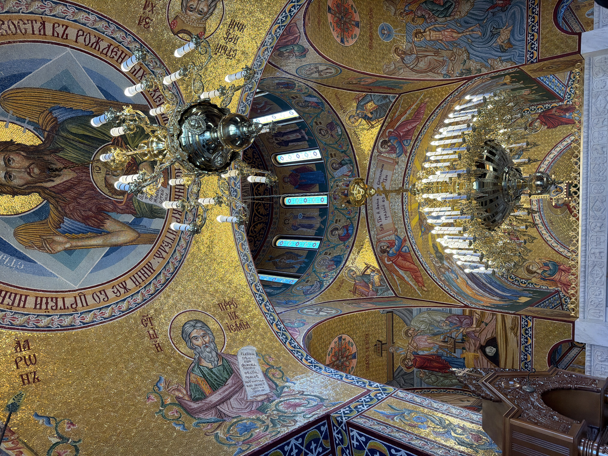 The image captures a view of the ornate, mosaic-covered ceiling of a church, likely located in Plovdiv, Bulgaria, given the presence of Cyrillic text. The ceiling is decorated with religious figures and symbols typical of Orthodox Christian iconography, rendered in gold and vibrant colors. Several large, ornate chandeliers with many lights hang from the ceiling, partially obscuring the mosaic details. A portion of a wooden structure, possibly a lectern or part of the church furniture, is visible in the foreground. The mosaics include depictions of angels, prophets, and scenes from the Bible. The scene is indoors, focusing on the architectural and artistic details of the church's interior.