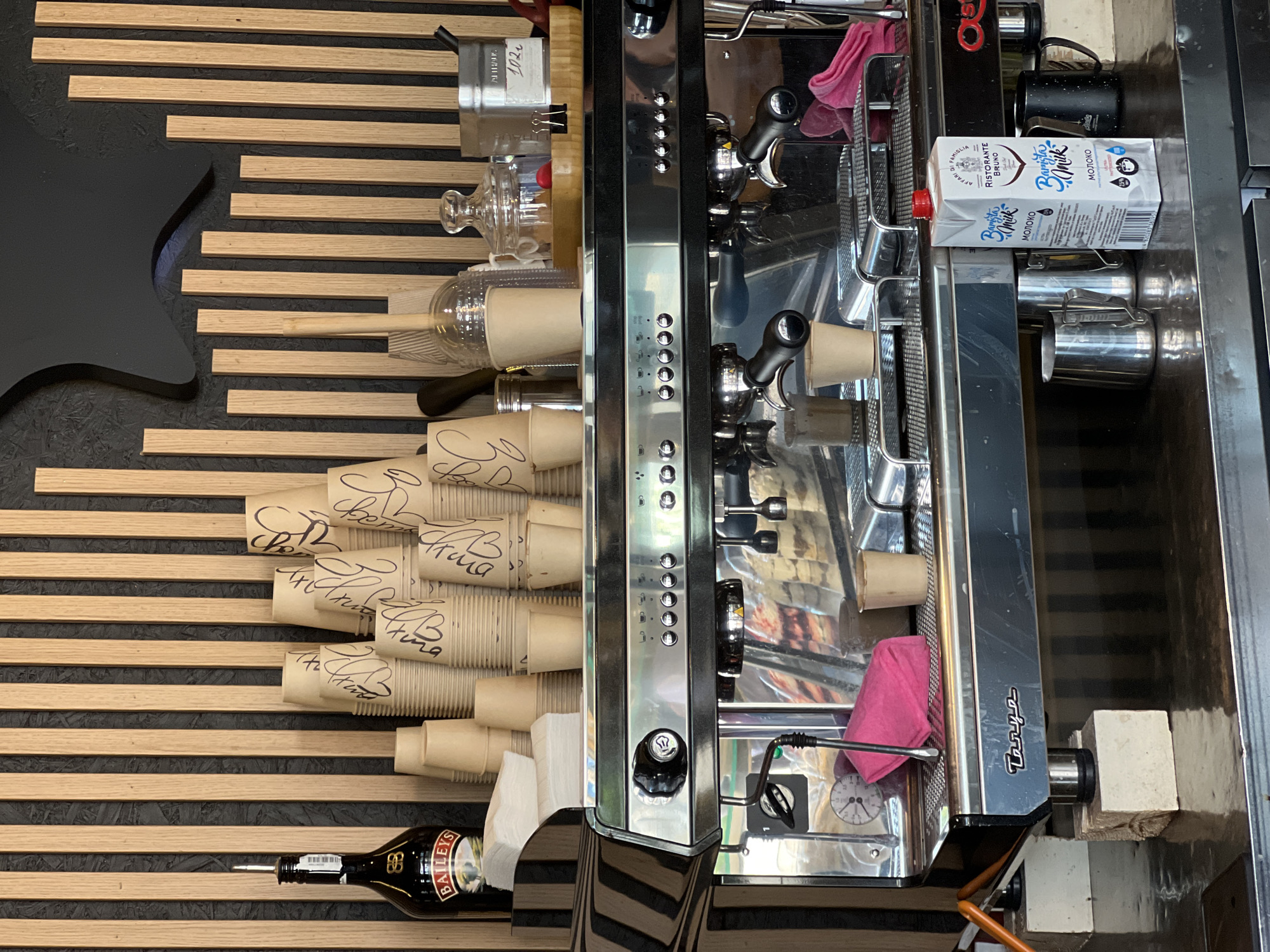 The image shows a counter in a cafe, likely in Fontanka, Ukraine, with a commercial espresso machine as the central focus. A bottle of Baileys liqueur stands to the left of a stack of paper cups. On top of the espresso machine are various coffee-related items, including more paper cups, a clear glass jar with a lid, and a wooden tray with items. The espresso machine itself is made of stainless steel and has multiple brew heads and control buttons. Below the machine, on the counter, are milk pitchers, a carton of "Barista Milk," and mugs. The background features a wall with vertical wooden slats. No people are visible in the scene.
