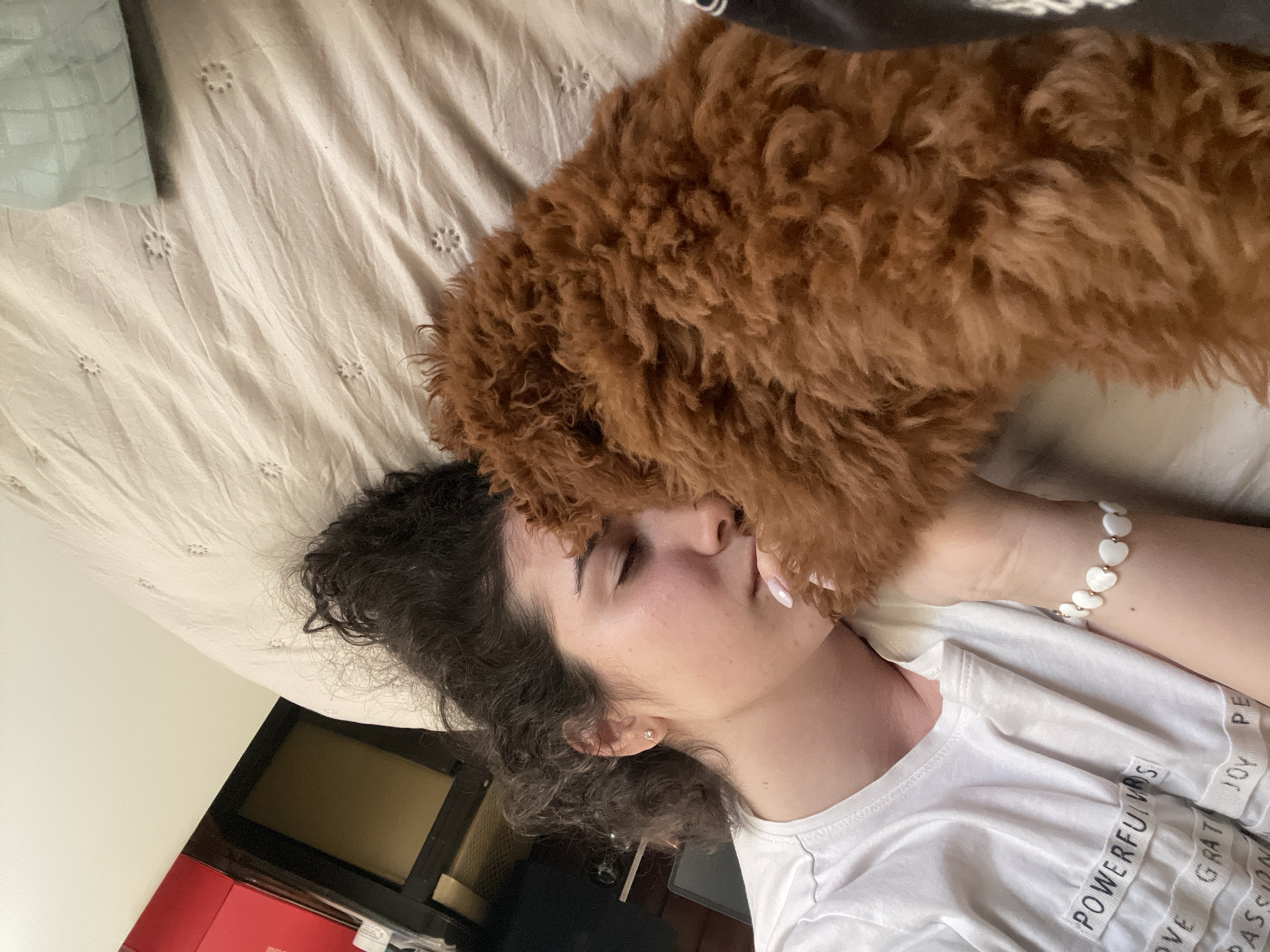 A woman with curly dark hair is lying on a bed with a light-colored embroidered bedsheet. She has her eyes closed and a small brown dog is resting its head on her face. The woman is wearing a white t-shirt with words printed on the sleeves, and a white heart bracelet on her wrist. Part of a light green pillow is visible in the upper left corner. The image is taken from a slightly elevated angle. There is no indication of the location being Sofia, Bulgaria.